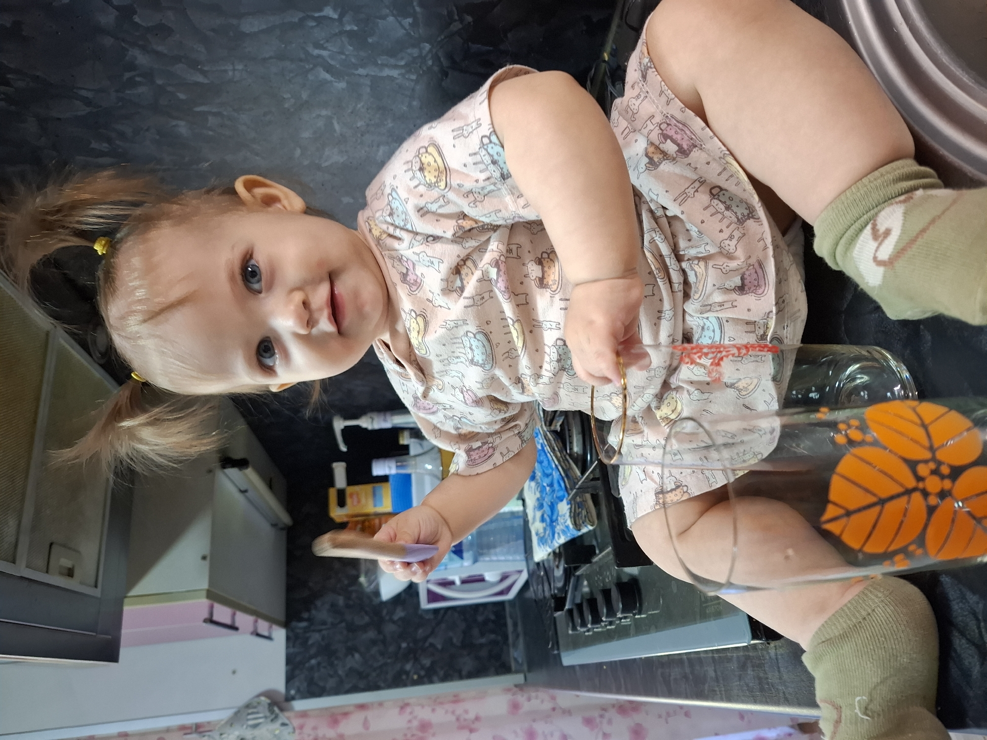 A curious toddler, likely a girl, with light brown hair styled in two pigtails with yellow ties, sits on a dark, possibly black, kitchen countertop. Her bright blue eyes are wide and focused on the camera, accompanied by a slight smile or smirk. She wears a light-colored, short-sleeved top or dress patterned with various cartoon animals and objects like teacups, and light green socks with a white and brown pattern.
In her right hand, she holds a small wooden brush with a purple-colored tip. Her left hand grasps a large, clear glass or vase adorned with a prominent orange, multi-leaf design. The background reveals a kitchen setting: a black stovetop with several burners and control knobs is visible to her right, and various bottles, dispensers, and containers are arranged on the counter behind her. Part of a metallic sink basin is seen to her left. Further back, light-colored kitchen cabinets with dark handles are visible, and the area below the counter features a light-colored, patterned surface. The child appears to be engaged in playful exploration of the kitchen items. This scene takes place indoors, likely during the daytime.
Location context: Chaikovsky, Russia.