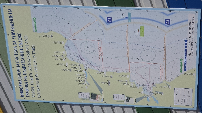 A large, vertically oriented informational sign is mounted against a corrugated metal building facade, displaying a detailed nautical chart of the Varna, Bulgaria, maritime area. The sign's title, presented in both Bulgarian and English, reads "ИНФОРМАЦИОННА СИСТЕМА ЗА УПРАВЛЕНИЕ НА ТРАФИКА НА ПЛАВАТЕЛНИТЕ СЪДОВЕ (VESSEL TRAFFIC MANAGEMENT AND INFORMATION SYSTEM (VTMIS))".
The chart, predominantly light blue for water and light beige/green for land, depicts the Black Sea coastline, Varna Bay, and navigational channels. It features multiple vessel icons, circular zones, designated areas labeled "A1", and various directional arrows (green and blue). Specific maritime infrastructure, depth markings, and textual annotations (primarily in Bulgarian) are visible throughout the map. The bottom portion of the sign displays "ДЪРЖАВНО ПРЕДПРИЯТИЕ "ПРИСТАНИЩНА ИНФРАСТРУКТУРА"" (State Enterprise "Port Infrastructure"). The sign itself is rectangular with a blue border and is secured to the building with visible fasteners. The background building consists of horizontal corrugated panels in varying shades of green, grey, and blue. No individuals or active interactions are present in the scene.