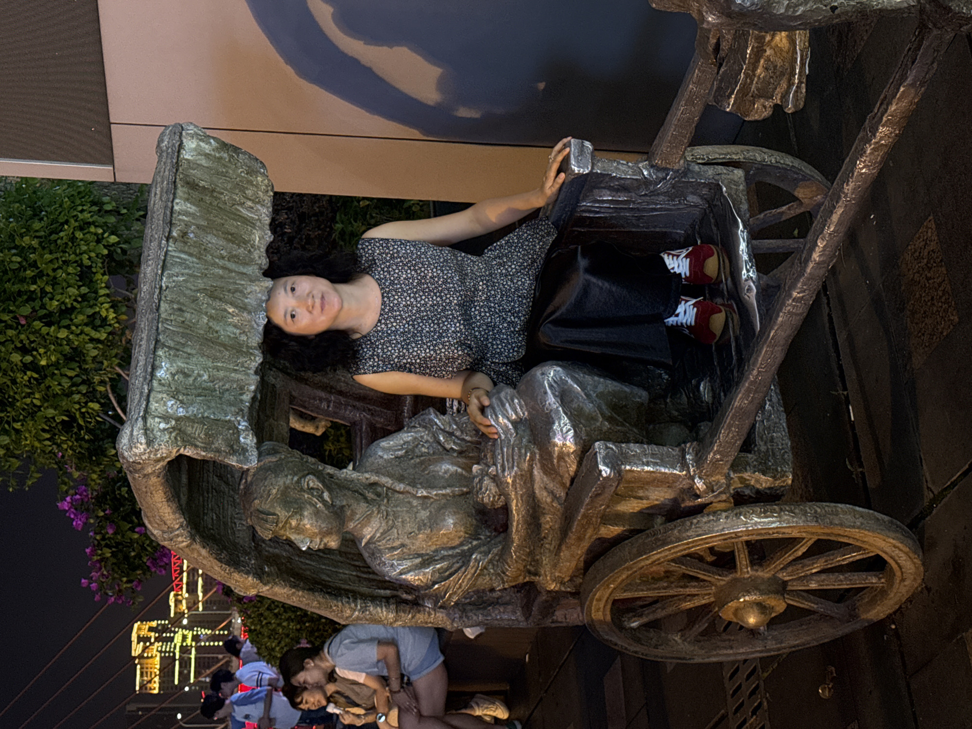 A woman is sitting in a large metal sculpture of a man pulling a cart in the Yuzhong District of China. The woman is wearing a patterned dress and black skirt, with red and white sneakers, and is holding onto the side of the cart. The sculpture is located near greenery, including bushes with purple flowers. In the background, lights from buildings can be seen at night, and other people appear to be in the vicinity.