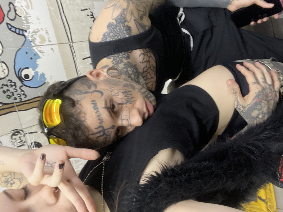 A tattooed man with sunglasses and a bandage on his face leans close to a woman. Both wear black clothing. The man's ear is gauged, and he has the word "MAMMA" tattooed on his forehead. Tattoos cover his hands, neck, and chest. The woman's hand is visible, and she also has tattoos. A black furry garment rests nearby. The background features a patterned surface.