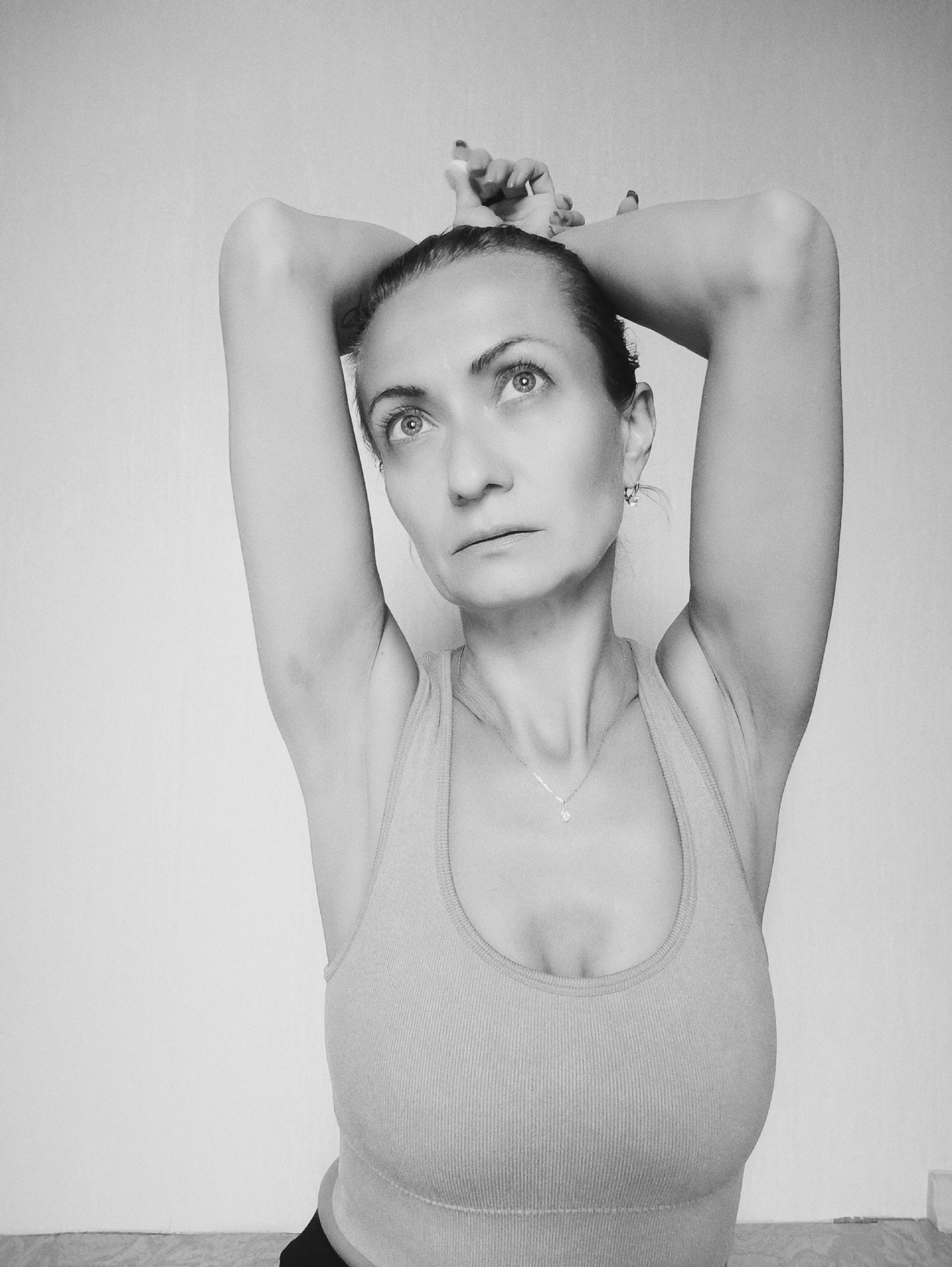 A black and white, waist-up, medium shot photograph shows a woman with her arms raised and hands clasped behind her head, looking upwards and to her left. She has dark hair pulled back and wears a light-colored, ribbed tank top with a thin necklace. The background is a plain, light-colored wall. There is no indication of the city of Moscow, Russia, or any other specific location.
