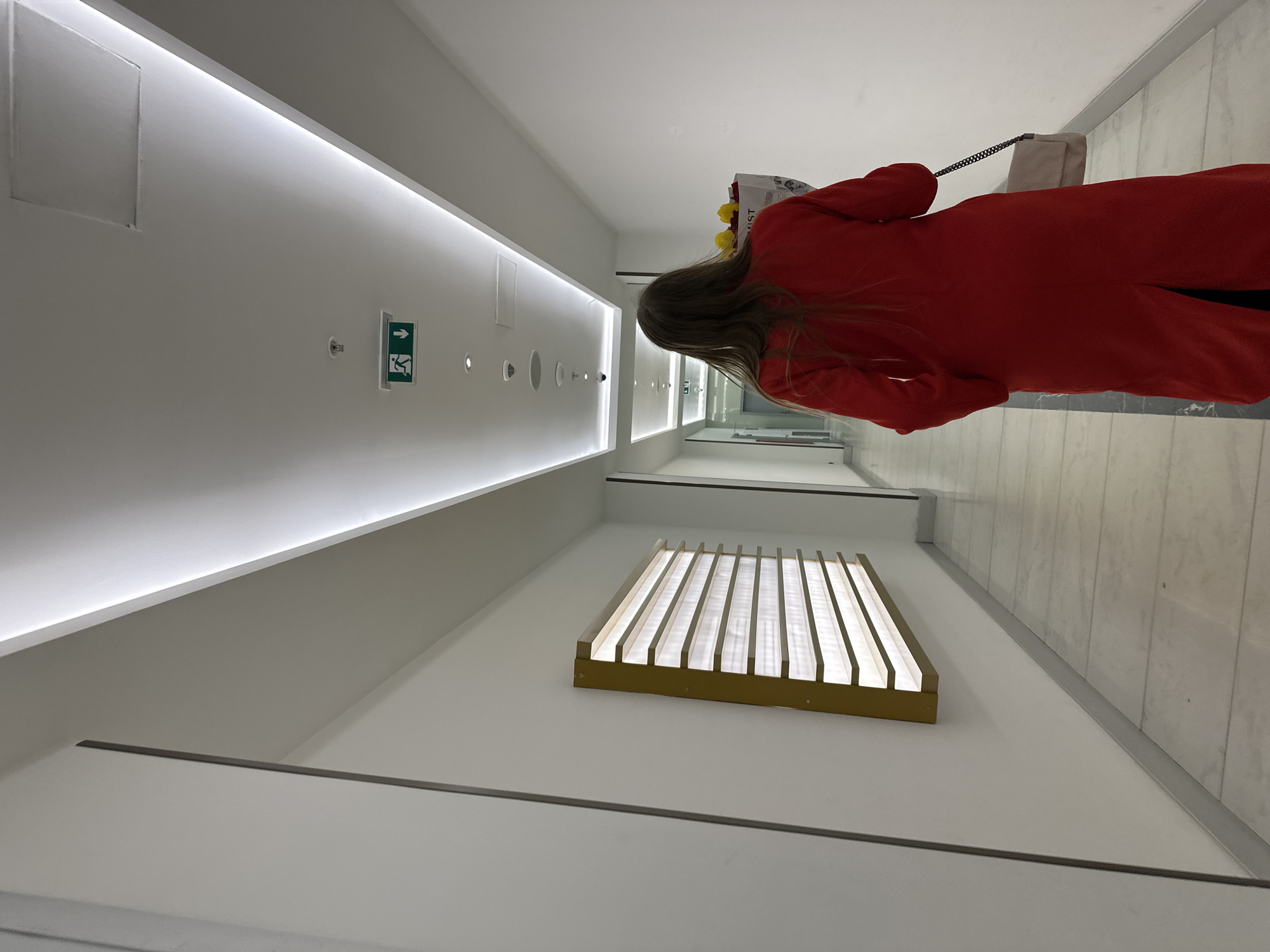 A woman wearing a red coat walks down a brightly lit hallway, possibly in a shopping center or commercial building. The hallway has a modern design with white walls, a light strip along the ceiling, and recessed lighting. An exit sign is visible on the wall. A window with slats is on the right. The location is likely in Batumi, Georgia, as that was mentioned as a location requirement.