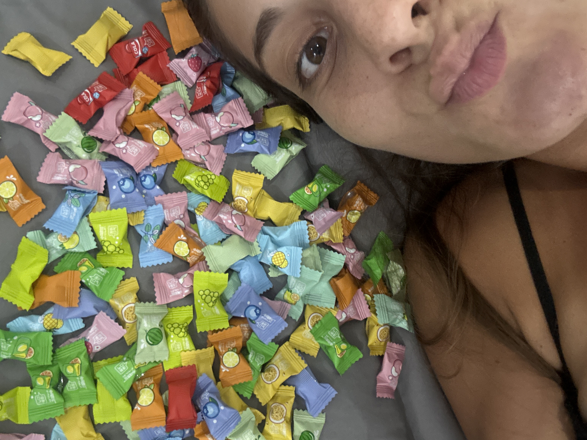 A selfie shows a woman lying on a gray surface, possibly a bed, surrounded by approximately 100 individually wrapped candies. The candies are in various colors including yellow, red, orange, pink, blue, and green, each appearing to have a small fruit image on the wrapper. The woman is positioned on the right side of the frame, partially obscuring the candies. Her face is close to the camera, showing one eye, part of her nose, and lips. A black strap of clothing, likely a bra or tank top, is visible on her right shoulder. No information in the image indicates this photo was taken in Đà Nẵng, Vietnam.