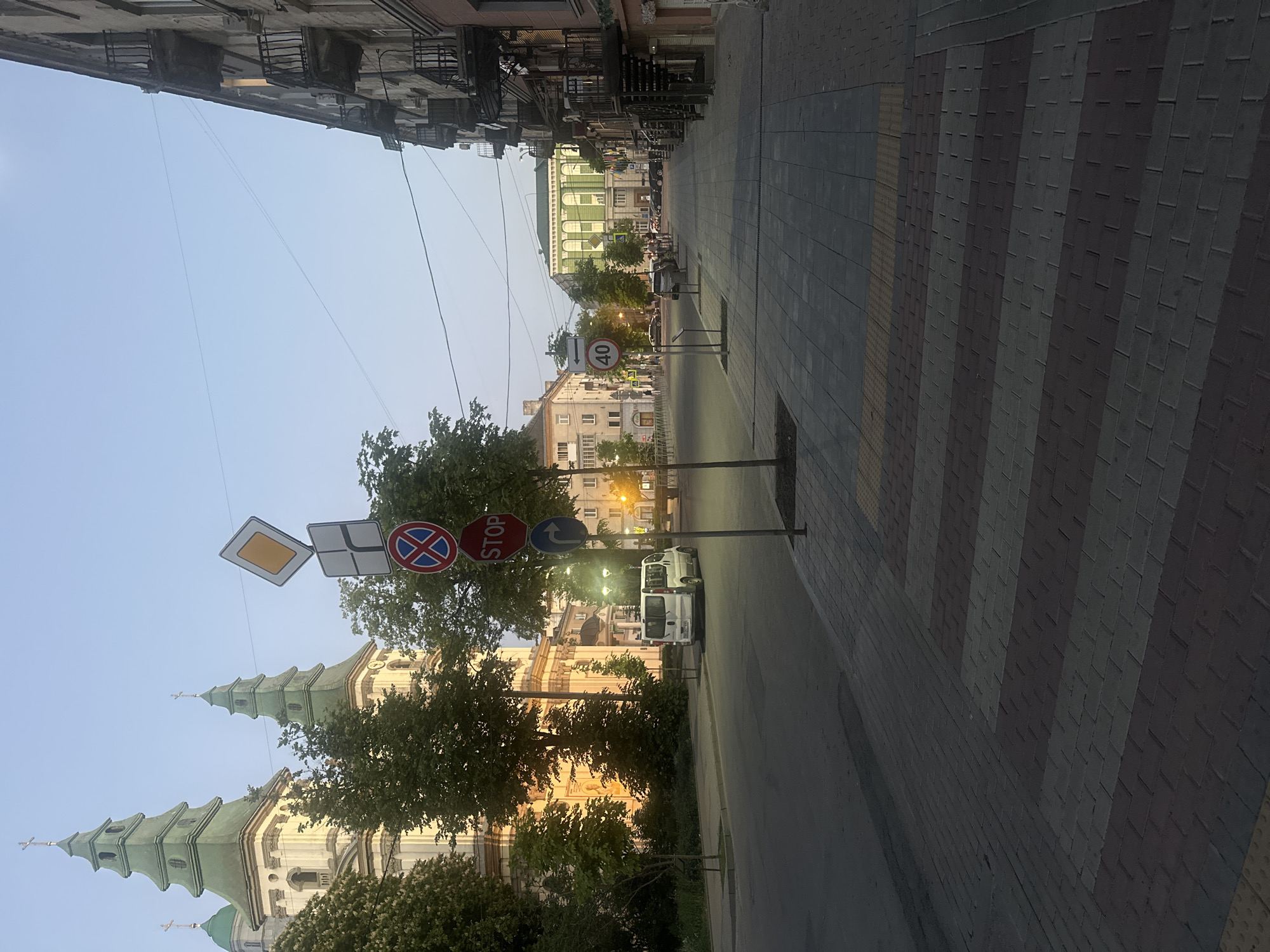 The image depicts a street in Ternopil, Ukraine. On the left, the spires of a church are visible behind trees, catching the late afternoon sun. Multiple road signs are clustered on a pole, including "STOP," "No Parking," and an advisory sign. The street itself is paved, with a striped brick pattern on the sidewalk. Buildings line both sides of the street, with architectural details like balconies and awnings. In the distance, a "40" speed limit sign is visible, along with parked cars. The sky is a clear, pale blue.
