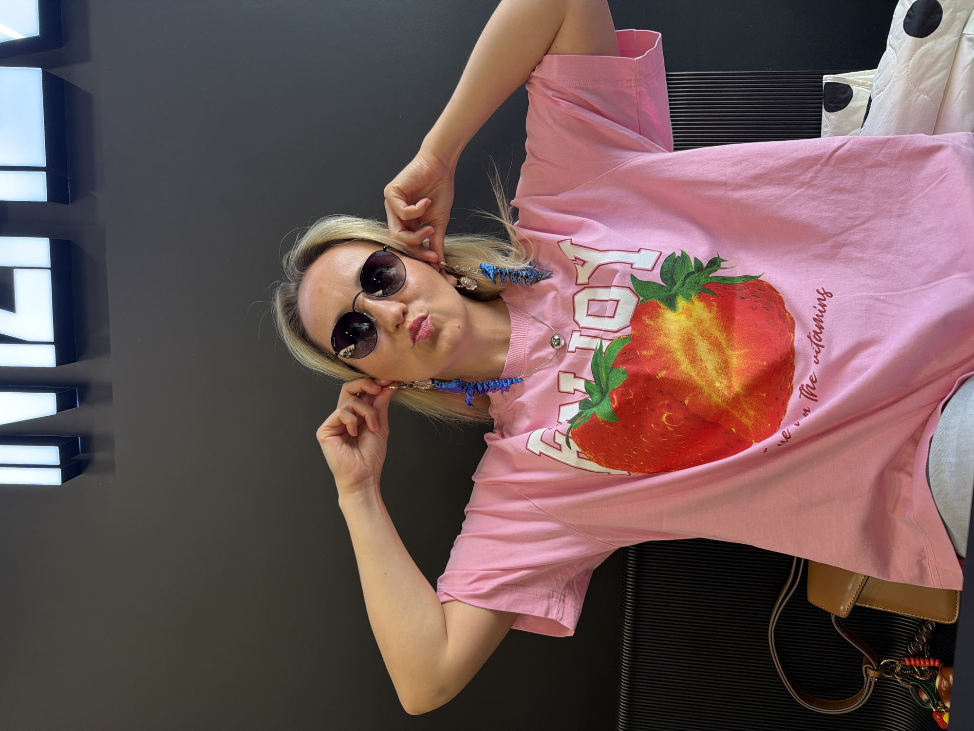 A woman with blonde hair and sunglasses is wearing a pink t-shirt with a strawberry graphic. She is holding her ears with her hands, and her mouth is puckered. The t-shirt has the words "ENJOY" and "it is the vitamins" printed on it. She is wearing blue earrings. There is a brown purse hanging from her shoulder. The background is a black wall with white illuminated letters spelling out "INTEX."