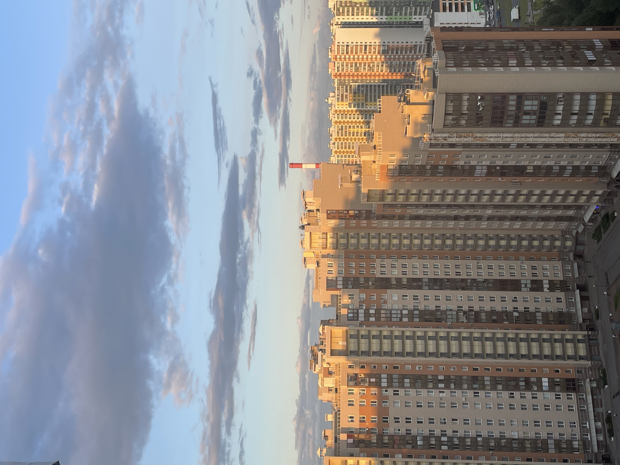 A high-angle, medium shot captures a dense urban skyline bathed in the warm, golden light of sunset. The sky above is a soft blue, adorned with scattered, wispy clouds that range in color from pale gray to tinges of pink and orange. In the distance, a tall, red and white striped chimney pierces the sky.
Dominating the foreground are numerous apartment buildings of varying heights and architectural styles. Many of these structures are tall, multi-story buildings with a repeating pattern of windows and balconies. The facades exhibit a mix of colors, including beige, light brown, white, and orange, with some buildings featuring darker brown or gray accents. The late afternoon sun casts long shadows, highlighting the texture of the buildings and creating a dramatic play of light and dark.
The lower portion of the image reveals the urban landscape below, featuring paved courtyards, patches of greenery, and some parked vehicles. The scene suggests a residential area within a large city, likely in Russia given the context, with the architecture typical of modern Russian urban development. No individuals are clearly discernible in the image.