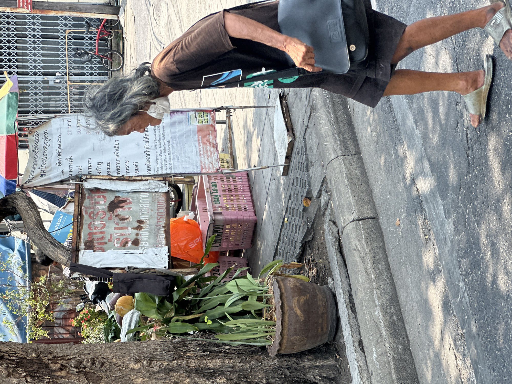 An older woman with gray hair, wearing a brown top, dark shorts, a face mask, and sandals, walks on a sidewalk in Bangkok, Thailand. She carries a dark-colored bag. The scene is at street level; a tree is on the left side of the photo. The woman walks past a street vendor stall featuring a price list sign, crates, and various items. A building with a red bicycle in front of it is in the background. The sign says "Klong Thom Wat Somanas" in Thai, indicating the street is near Klong Thom and Wat Somanas Temple. The sign lists taxi fare prices.