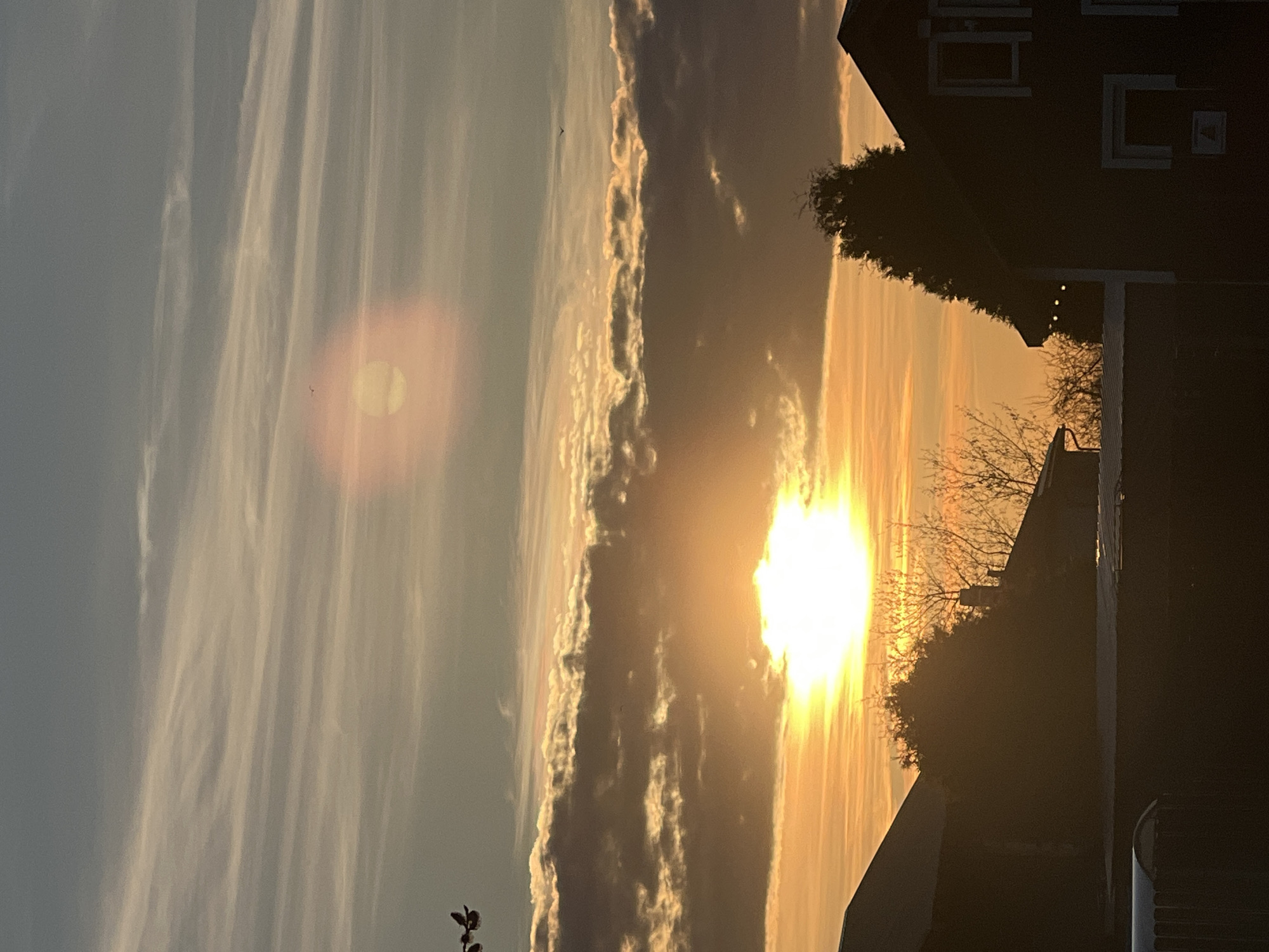 The image depicts a sunset scene, likely in an urban or suburban environment, based on the presence of buildings in silhouette. The sun is the primary light source, creating a bright, golden glow that dominates the sky. Wispy clouds are visible, partially obscuring the sun and causing a lens flare effect to the left of the sun. The buildings are silhouetted against the sky, including residential structures with visible windows. The setting suggests a location where urban development meets a natural setting with unobstructed sky views. The specific location cannot be determined with the provided image. There is no evidence of people, subjects, objects, actions, or interactions. The environment can be described as urban meets nature. There are no clues about Riga or Latvia in this photo.