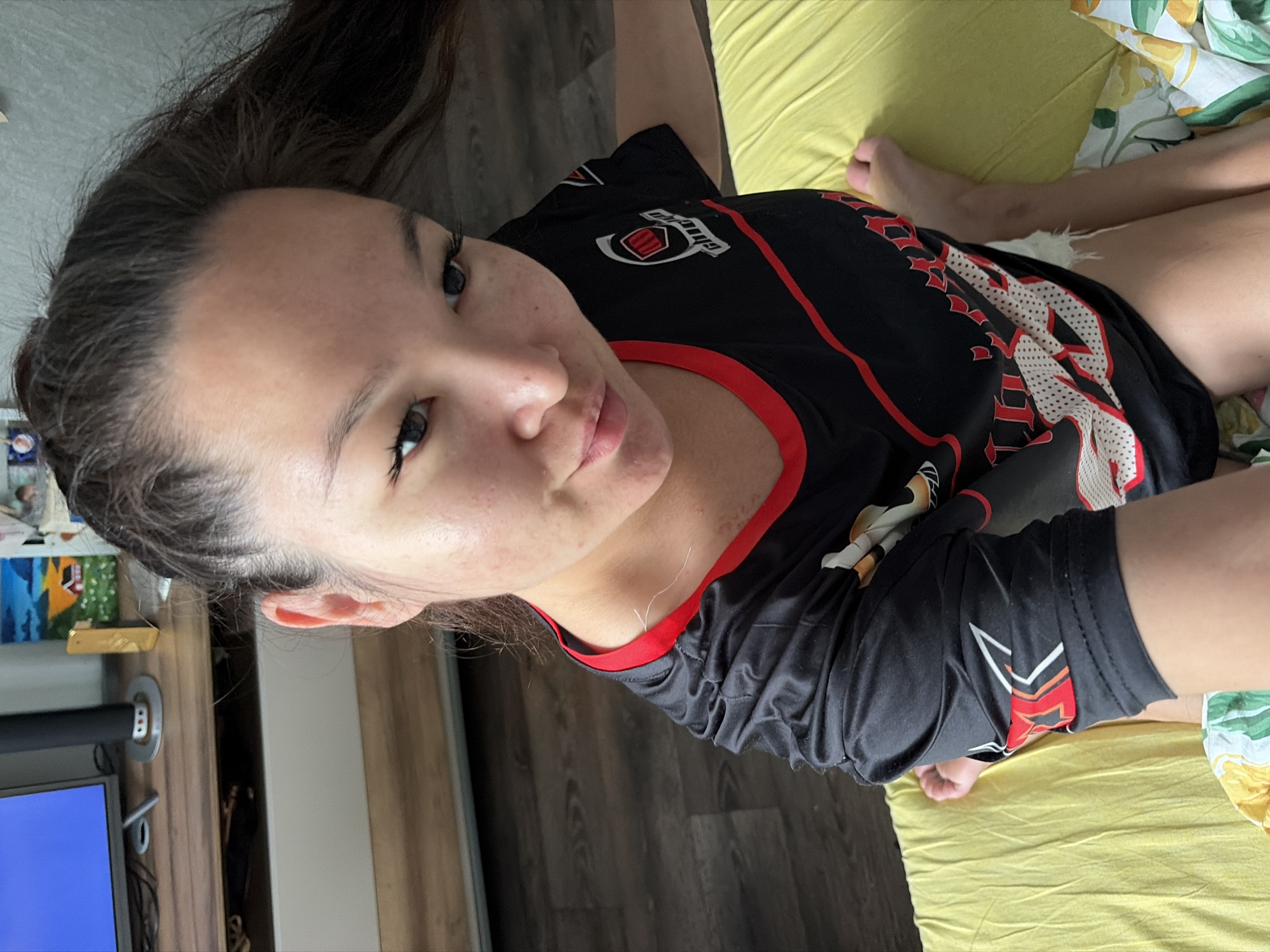 A woman with dark hair is sitting on a yellow bedspread, her body angled towards the camera. She is wearing a black t-shirt with red trim and a logo on the left chest. Her hair is pulled back and to the right, partially obscuring her face from a top-down view. The background shows a wooden floor and a piece of furniture with a television and decorative items. This image was taken in Chaikovsky, Russia.