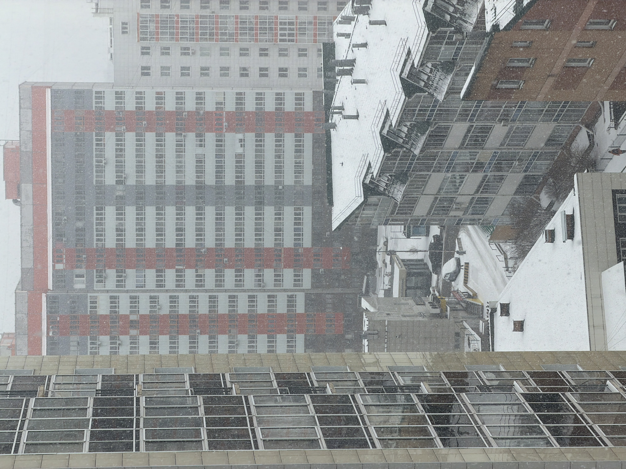 The image depicts an urban landscape in Krasnoyarsk, Russia, observed from an elevated viewpoint during a heavy snowfall. The scene is dominated by multiple architectural structures under an overcast, daytime sky.
In the foreground, the roof of a large, modern building features an intricate grid of glass panels, resembling a skylight system, partially covered in snow and precipitation. Beyond this, a prominent multi-story building occupies the left midground, distinguished by its facade with alternating grey and white sections broken by horizontal red stripes. This building is densely punctuated with rectangular windows and some external air conditioning units.
To the right of the striped building, a complex of other structures is visible. One building exhibits a reddish-brown, brick-like exterior with dark, rectangular windows. Adjacent to it, another building presents a unique multi-tiered, angular roof design, heavily blanketed in white snow. Further in the background, more buildings are present but are less distinct due to the falling snow and distance.
The prevailing weather is a heavy snowfall, with numerous snowflakes clearly visible against the buildings and in the air, contributing to a muted, soft atmosphere. Snow has accumulated on rooftops and visible ground surfaces. No specific human activity is clearly discernible, nor is any visible text present. The overall impression is one of a cold, quiet winter day in the city.