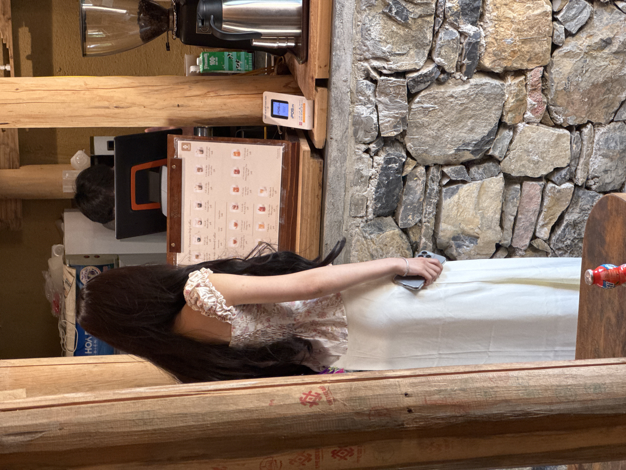 The image depicts a scene at a café in Lũng Cú Commune, Vietnam. A person with long, dark hair wearing a floral-patterned top and a white skirt is seated at a wooden railing. The person is holding a smartphone in their hand. Behind them, an indoor café space is visible, with coffee-making equipment and a menu board with Vietnamese writing. The wall behind the café structure is made of rough stone.