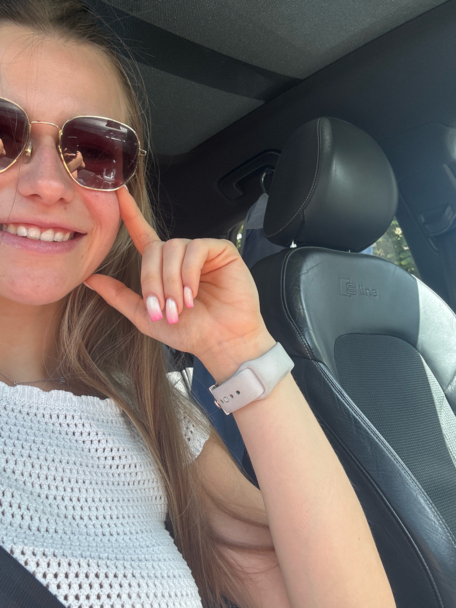 In the foreground, a young woman with blonde hair and a white crocheted top is sitting in the driver's seat of a car. She is wearing aviator sunglasses with gold frames and her left hand is touching her cheek. Her fingernails are painted with a pink and white ombre design. A light gray smartwatch is visible on her left wrist. The car's interior features black leather seats with the "S line" logo embossed on them. The background shows a glimpse of trees and sunlight, suggesting an outdoor location. The woman is likely in a car parked or driving, possibly in a city or a suburban area. The location in the city of Sozopol, Bulgaria is not evident from the image itself.