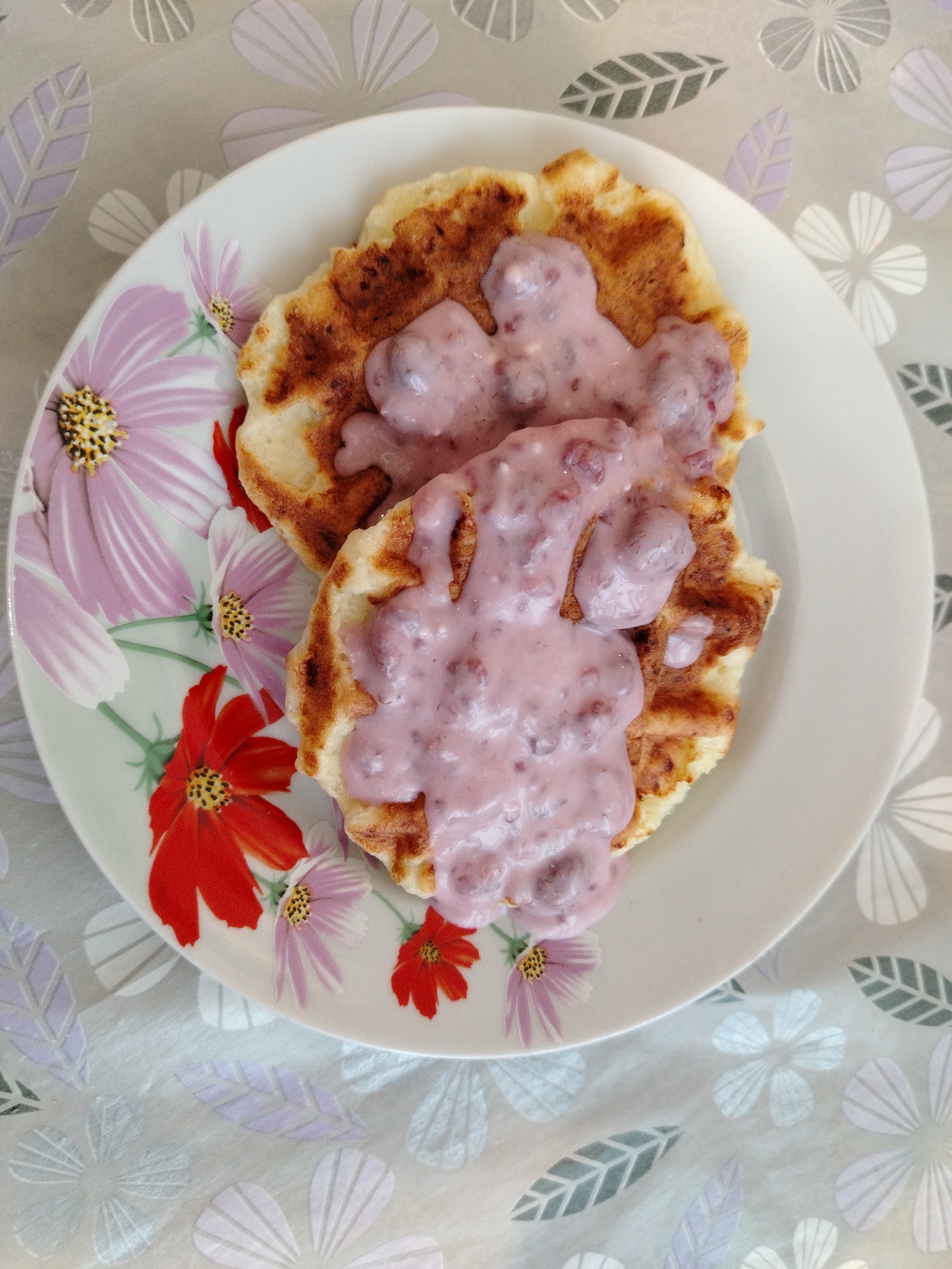 A top-down, full shot shows two irregularly shaped pancakes, stacked, on a white plate decorated with red and purple flowers. The pancakes are covered in a pink, creamy sauce that contains small pieces of fruit. The plate sits on a tablecloth patterned with white, lavender, and gray stylized flowers and leaves. The pancakes appear to be browned. The lighting is bright and even. No people are visible in the image. The image is described as taken in Chaikovsky, Russia, but no other details confirm that location.