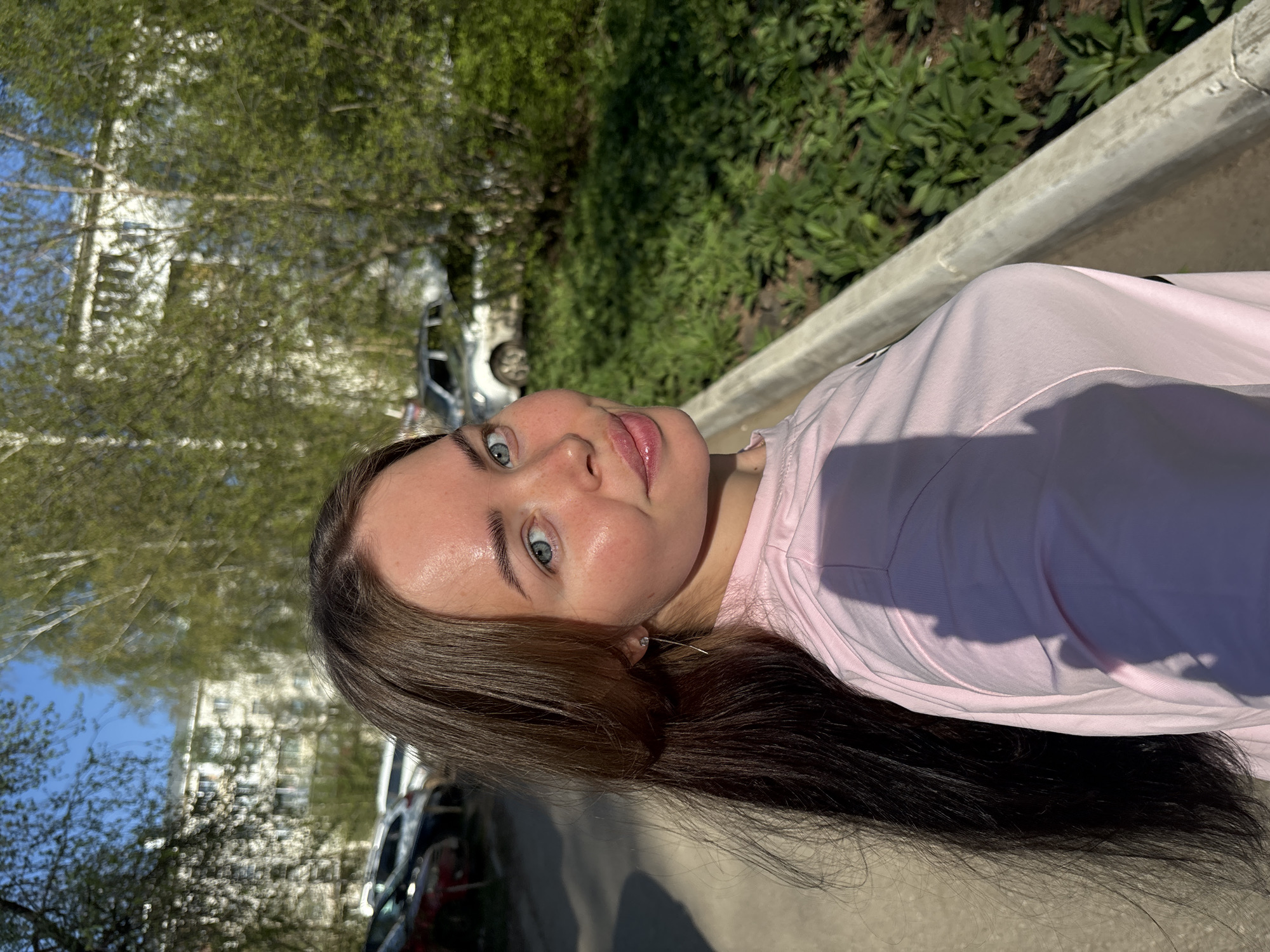 A young woman with long brown hair and blue eyes is the central subject of this outdoor portrait. She is wearing a light pink top and appears to be looking directly at the camera. The background reveals a street scene in what is likely Izhevsk, Russia, as evidenced by architectural styles. There is a mix of greenery, including trees and bushes, along with a section of concrete pavement or sidewalk. Vehicles are visible in the background, suggesting a residential or commercial area. The lighting indicates it is likely a sunny day with shadows cast on the subject and the surrounding environment.