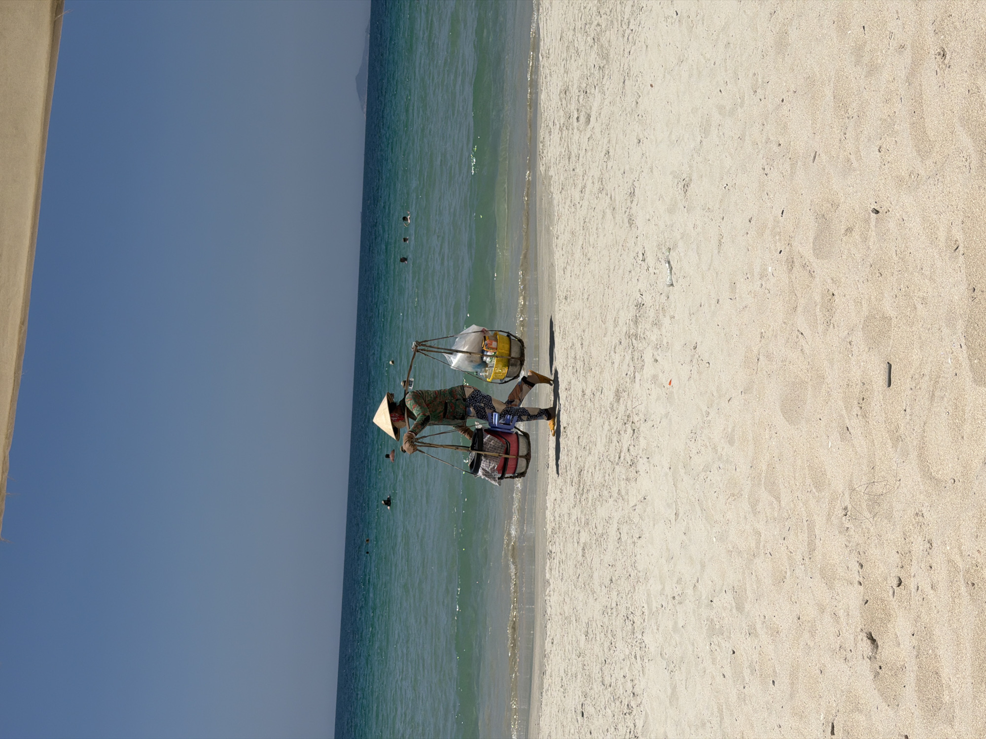 A forensic examination of the image reveals a sunlit coastal scene in Dong Ninh Hoa Ward, Vietnam. The primary subject is an individual traversing a light-colored sandy beach from left to right, positioned approximately one-third up from the bottom of the frame. This individual wears a conical straw hat (nón lá), a long-sleeved patterned garment, dark patterned trousers, and yellow closed-toe shoes. They are actively engaged in carrying a shoulder pole (gánh), from which two containers are suspended: a dark-colored basket with a red fabric element and a small blue stool attached on the left, and a yellow container filled with various items, some enclosed in clear plastic bags, on the right.
The beach itself is characterized by fine, light-colored sand with subtle indentations and minimal scattered debris, including small organic matter. The water's edge exhibits gentle, clear turquoise waves lapping against the shore.
In the mid-ground, the ocean is calm, transitioning from a turquoise hue near the shore to a deeper blue further out. Several individuals, identifiable by their heads and upper bodies, are visible swimming or wading in the ocean. On the far right horizon, a faint silhouette of a distant landmass, likely a hill or island, is discernible.
The sky occupies the upper portion of the image, presenting a clear, uninterrupted expanse of light blue. The top edge of the frame reveals a partial view of a light-colored, indeterminate structure, possibly a canopy or umbrella. Bright daylight illuminates the entire scene, indicating fair weather conditions.