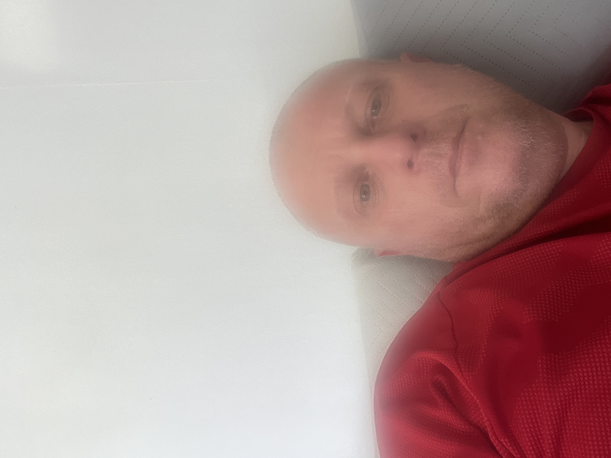 A bald man with light skin is lying down. He is wearing a red shirt. The background consists of a white wall and a patterned headboard or pillow. The location is unspecified in the city of Basingstoke and Deane, United Kingdom.