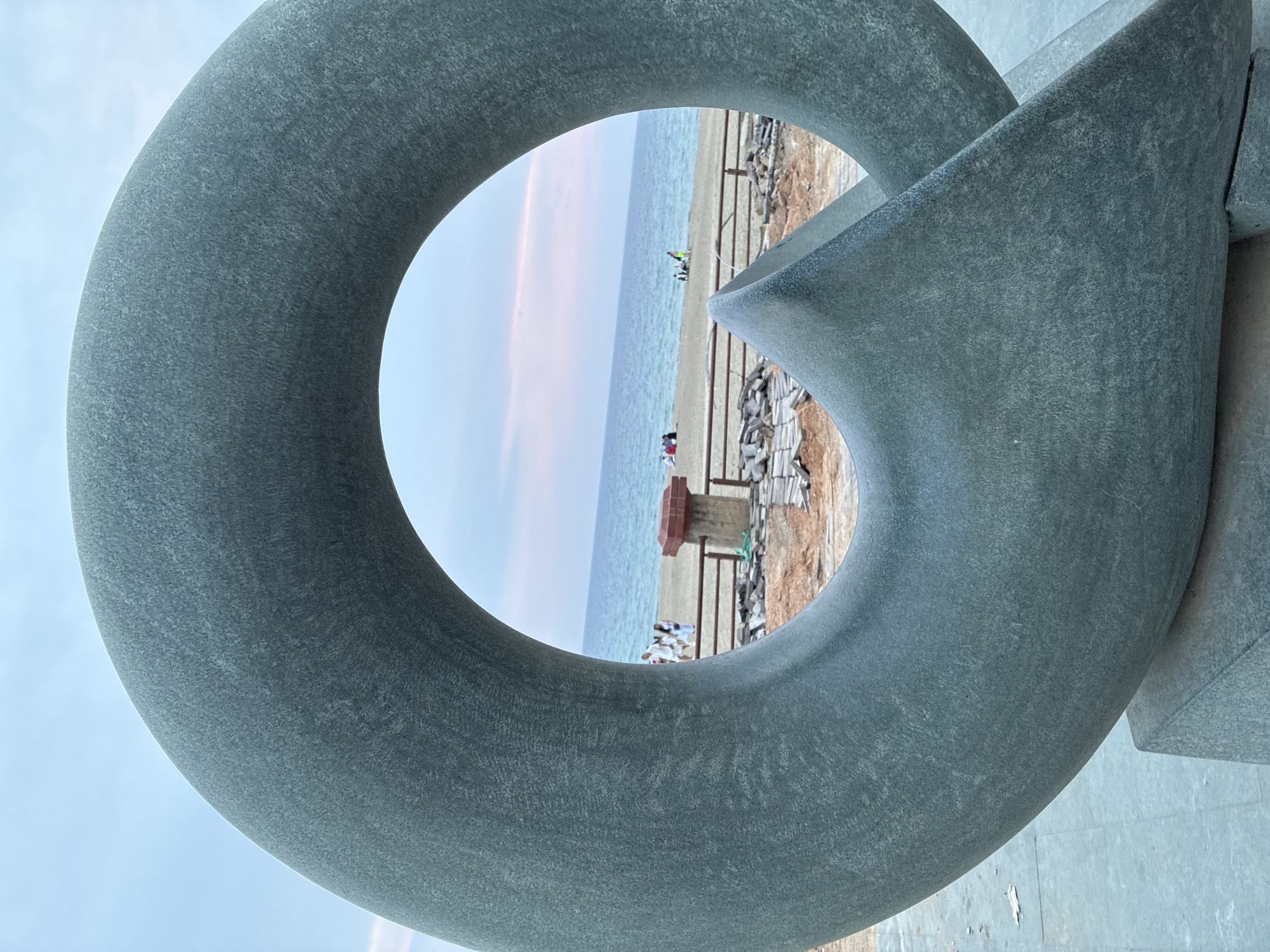 The image shows a close-up of a large, grey, stone sculpture with a circular opening. Through the opening, a beach and the sea are visible. The sky is overcast with some pink hues, suggesting either dawn or dusk. In the background, on the beach, a few people are scattered, some sitting and others standing. A wooden fence runs along the edge of the beach, separating it from an area with construction materials. The sculpture is situated on a paved area. The location is identified as the Cleopatra Beach in Alanya, Turkey, near a sculpture by the artist Yavuz Çiftçioğlu.