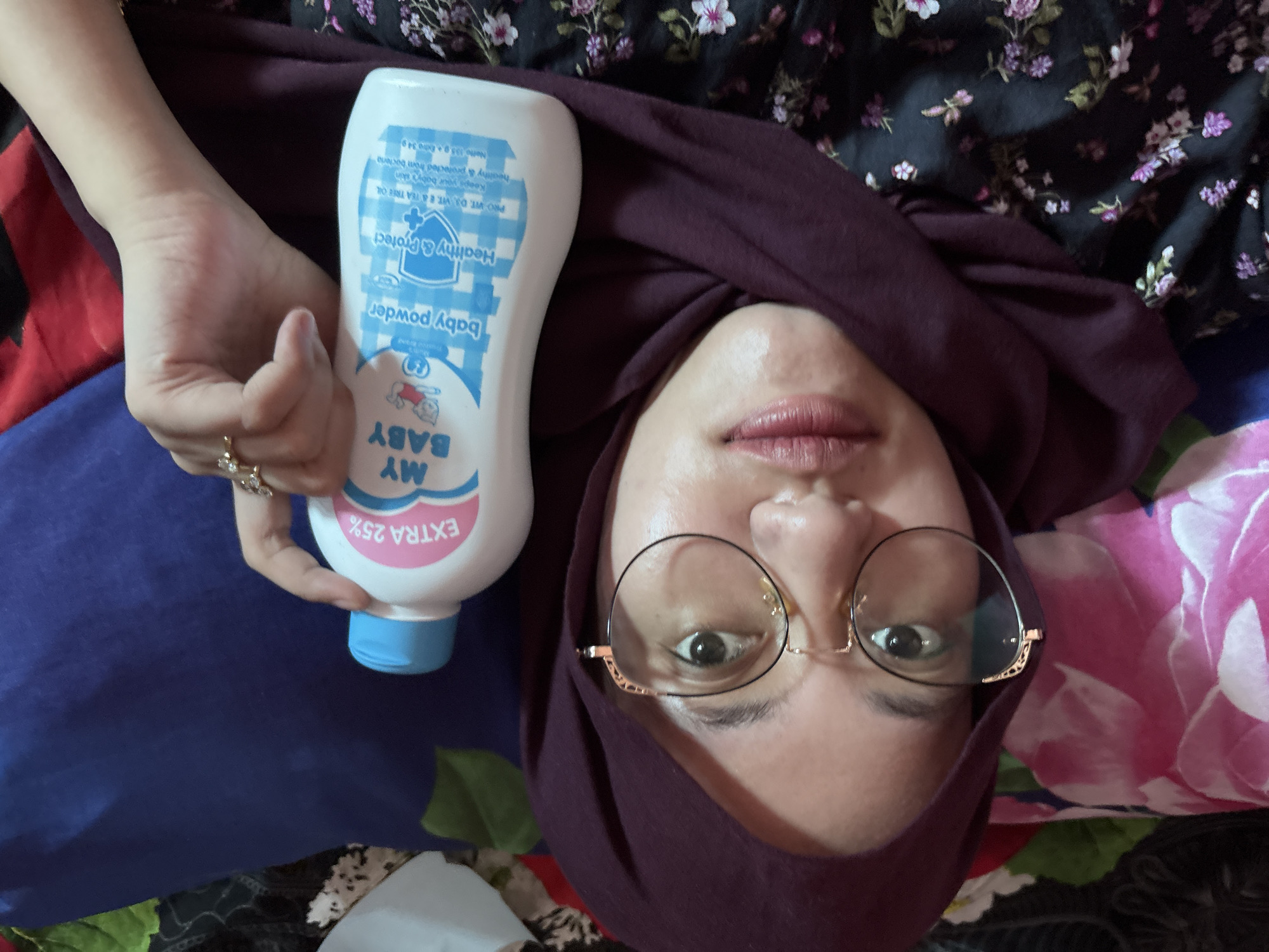 A woman wearing glasses and a purple hijab is holding a bottle of "My Baby" baby powder in her left hand. She is lying down, looking at the camera. The background includes a floral patterned fabric and a blue fabric. The text on the baby powder bottle includes "EXTRA 25%" and "Healthy & Protect". The location is Gadingrejo, Indonesia.