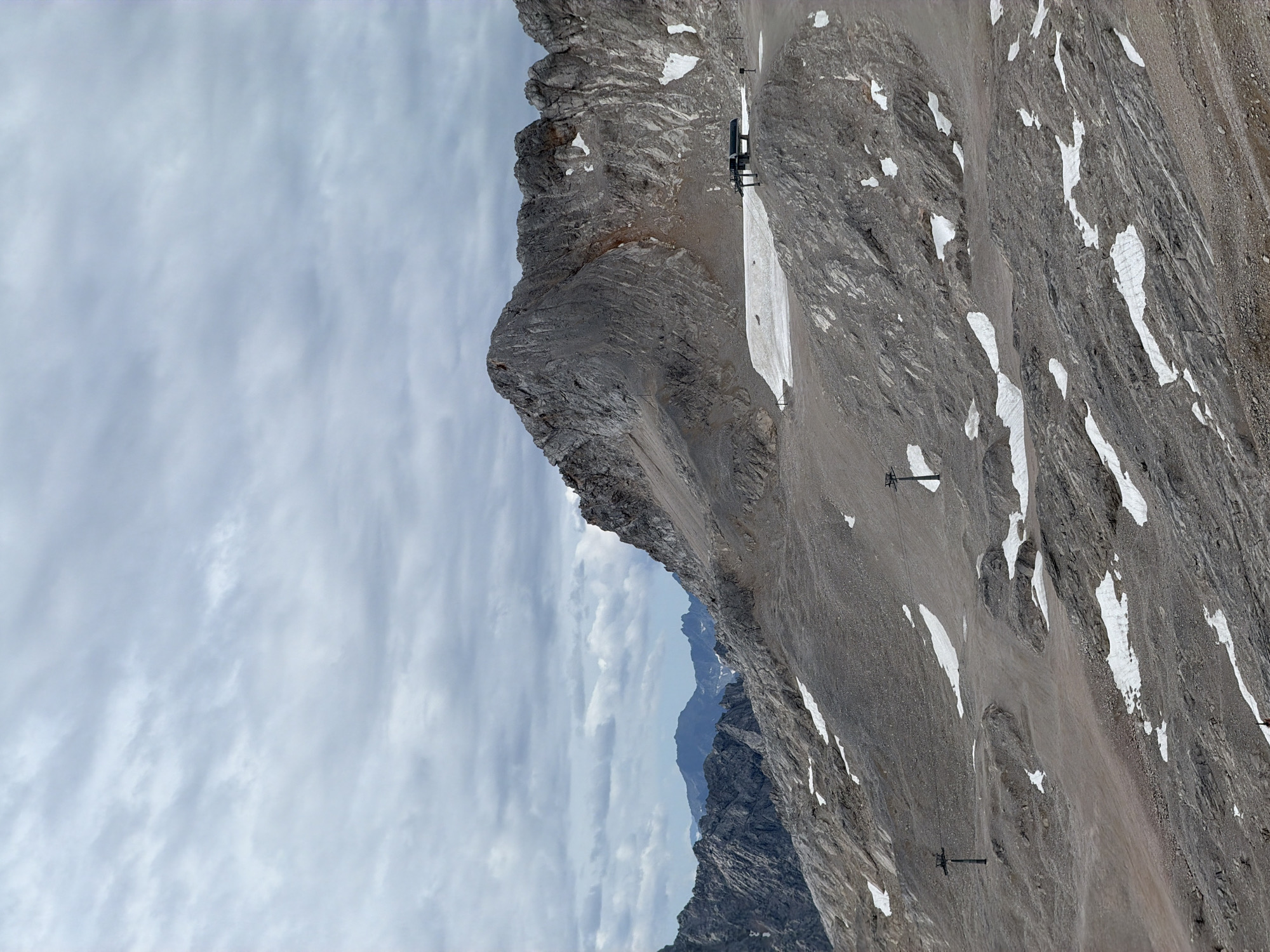The image depicts a mountainous landscape with a cloudy sky. The terrain is rocky and barren, with patches of snow scattered across the slopes. In the mid-ground, there is a ski lift station with a chairlift. Further into the distance, several mountain peaks are visible. The scene is likely a high-altitude location in the Alps, possibly near Garmisch-Partenkirchen, Germany, known for its alpine scenery and winter sports.