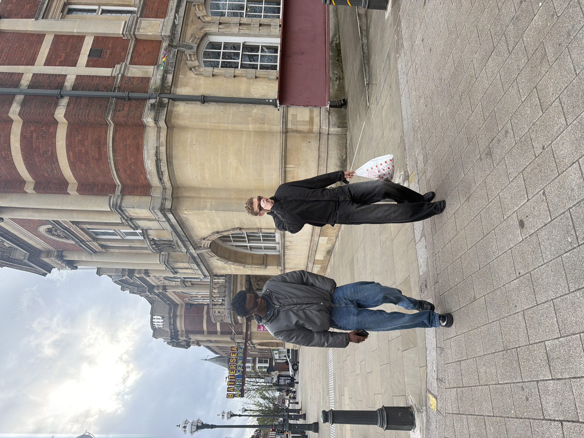 A medium shot of two men walking on a brick sidewalk in front of a building. The image is oriented sideways.
The first man is wearing a black jacket, black pants, and black shoes. He is holding a white bag with red polka dots. He is also wearing sunglasses or a mask.
The second man is wearing a grey jacket, blue jeans, and black shoes. He is holding a cell phone in his hand.
The building behind them is made of brick and stone. There is a sign that says "Battersea Arts Centre" on the building. There are also street lights in front of the building. The sky is cloudy. The location is Battersea, London, United Kingdom.