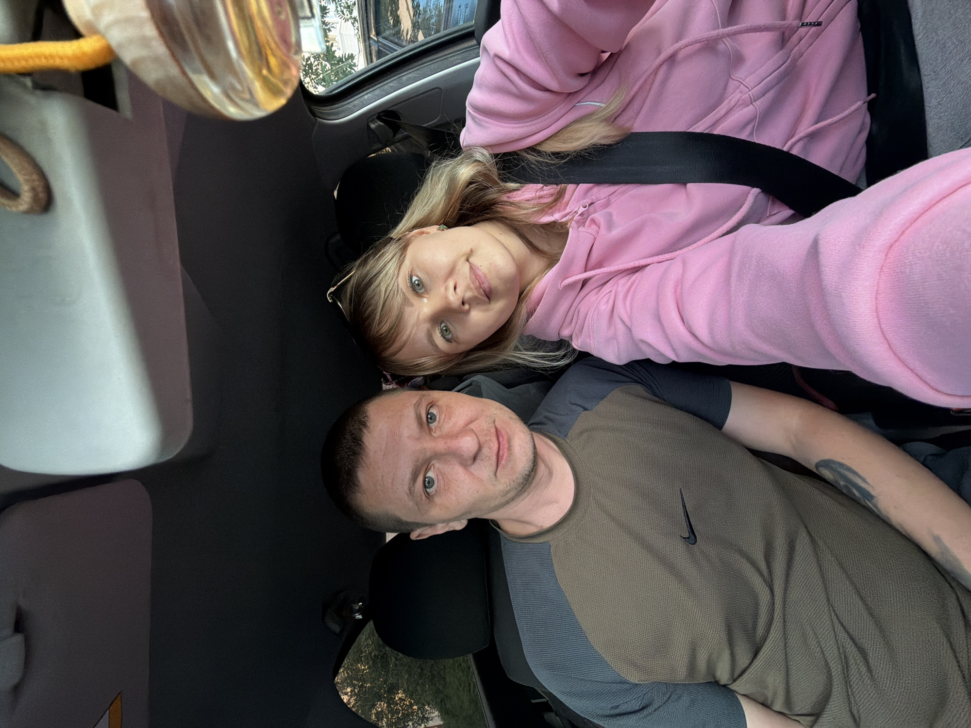 The image shows a couple in the front seats of a car, likely parked on a street in the city of Chaikovsky, Russia. The man, on the left, has short brown hair, blue eyes, and is wearing a two-toned t-shirt with a Nike logo. He has a tattoo on his left forearm. The woman, on the right, has blonde hair and is wearing a pink hooded jacket. Both individuals are looking at the camera, suggesting they are taking a selfie. The car's interior is visible, including the dashboard, sun visors, and seatbelts. A car air freshener hangs from the rearview mirror. Outside the car, trees and buildings can be seen through the windows, indicating an urban or suburban setting. The lighting suggests it is daytime, possibly late afternoon given the warm tones.