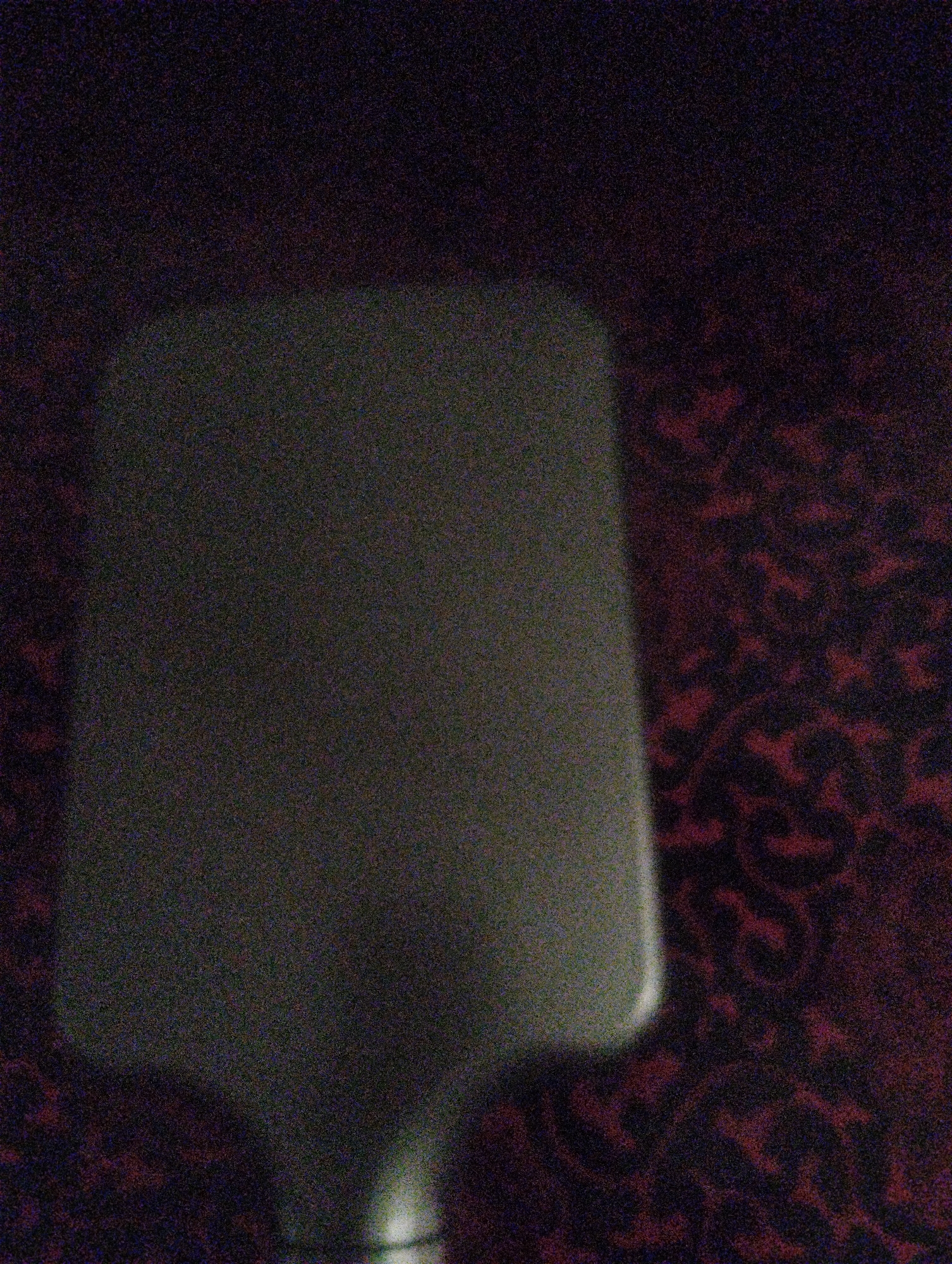 A dimly lit, grainy image shows a light-colored object, possibly a utensil or serving tool, angled diagonally across the frame, obscuring a patterned surface. The background is a dark, reddish-brown fabric with an ornate, repeating pattern. The lighting is poor, creating significant noise in the image. There are no discernible people or specific urban elements to indicate a city or location.