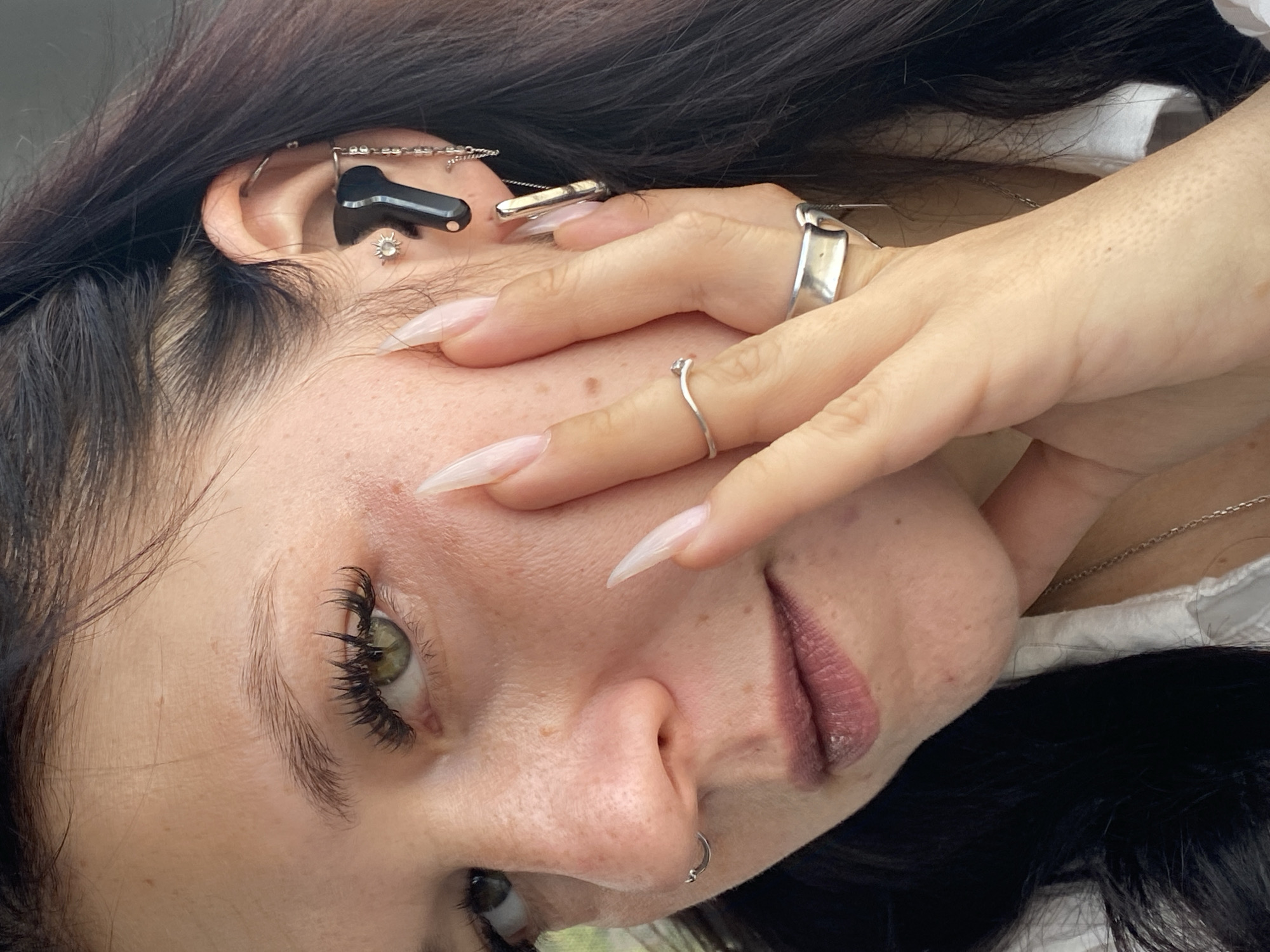 A close-up, eye-level, outdoor, natural light, medium-shot photograph captures a person's face and upper body. The subject, positioned slightly off-center to the left, has dark hair, green eyes, and is wearing dramatic false eyelashes, noticeable freckles on their cheeks and nose, and a nose ring on their left nostril. Their right hand, with long, acrylic nails, is brought up to their cheek, with fingers resting against their face. They are wearing several rings on their right hand, including a thin, wavy silver ring on the index finger and a wider, flat silver ring on the middle finger. In the subject's right ear, they are wearing multiple earrings, including a small sun-shaped stud, a black wireless earbud, and several delicate chain earrings. The subject is also wearing a white collared shirt and a silver necklace. The background is blurred, suggesting an outdoor setting, possibly in a park or garden, with hints of green foliage. The overall impression is one of a posed portrait, likely taken in Saint Petersburg, Russia, given the provided location context, though no specific landmarks are identifiable.