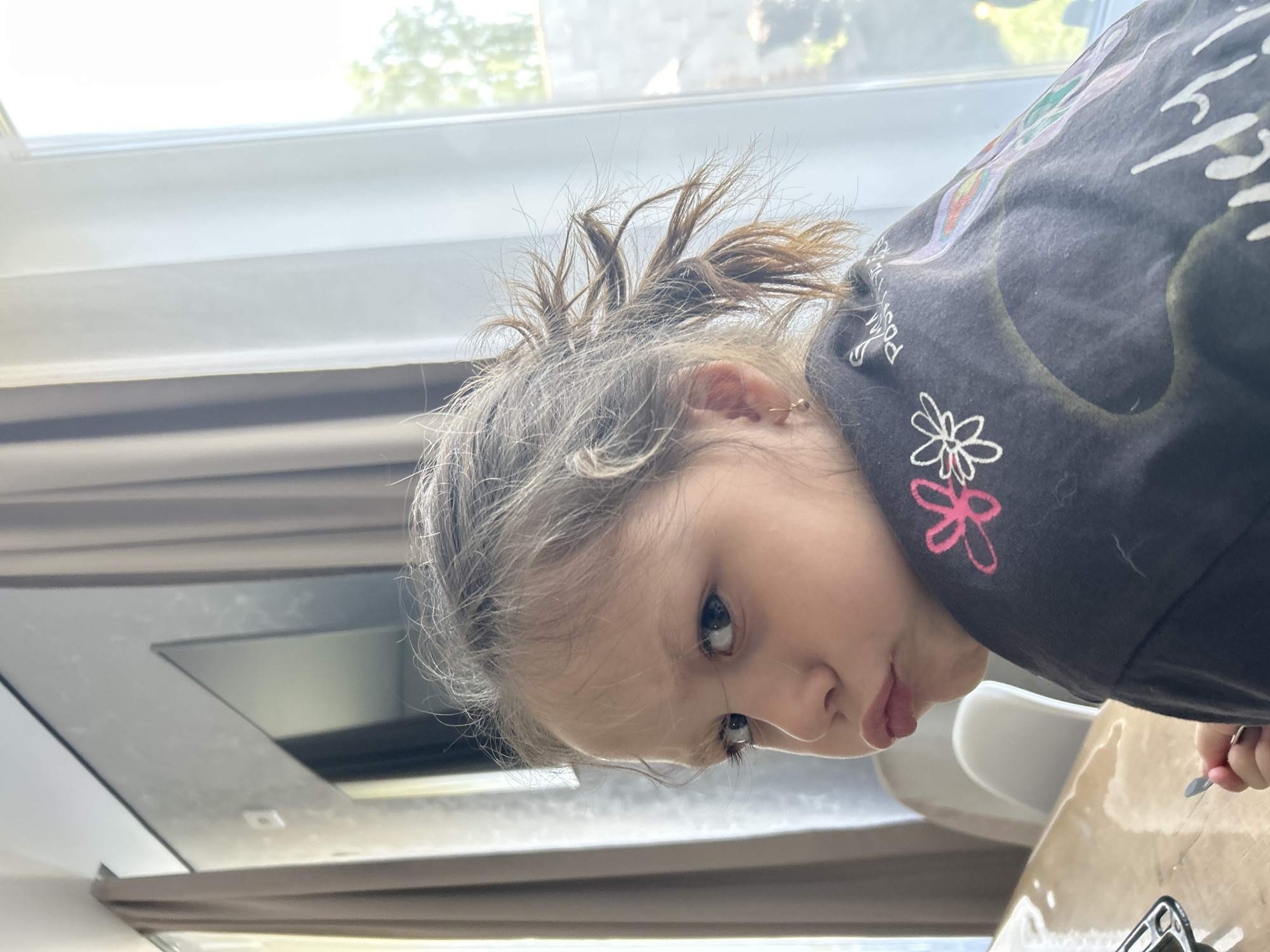 A young girl with brown and gray hair is looking down at something on a table. She is wearing a dark colored shirt with floral designs and a white and pink flower is visible on the shoulder. She has her hair pulled back into a small ponytail. The girl is located in what appears to be a home with natural light coming in from a window. The location in the city of Chaikovsky, Russia is not discernible in the image.
