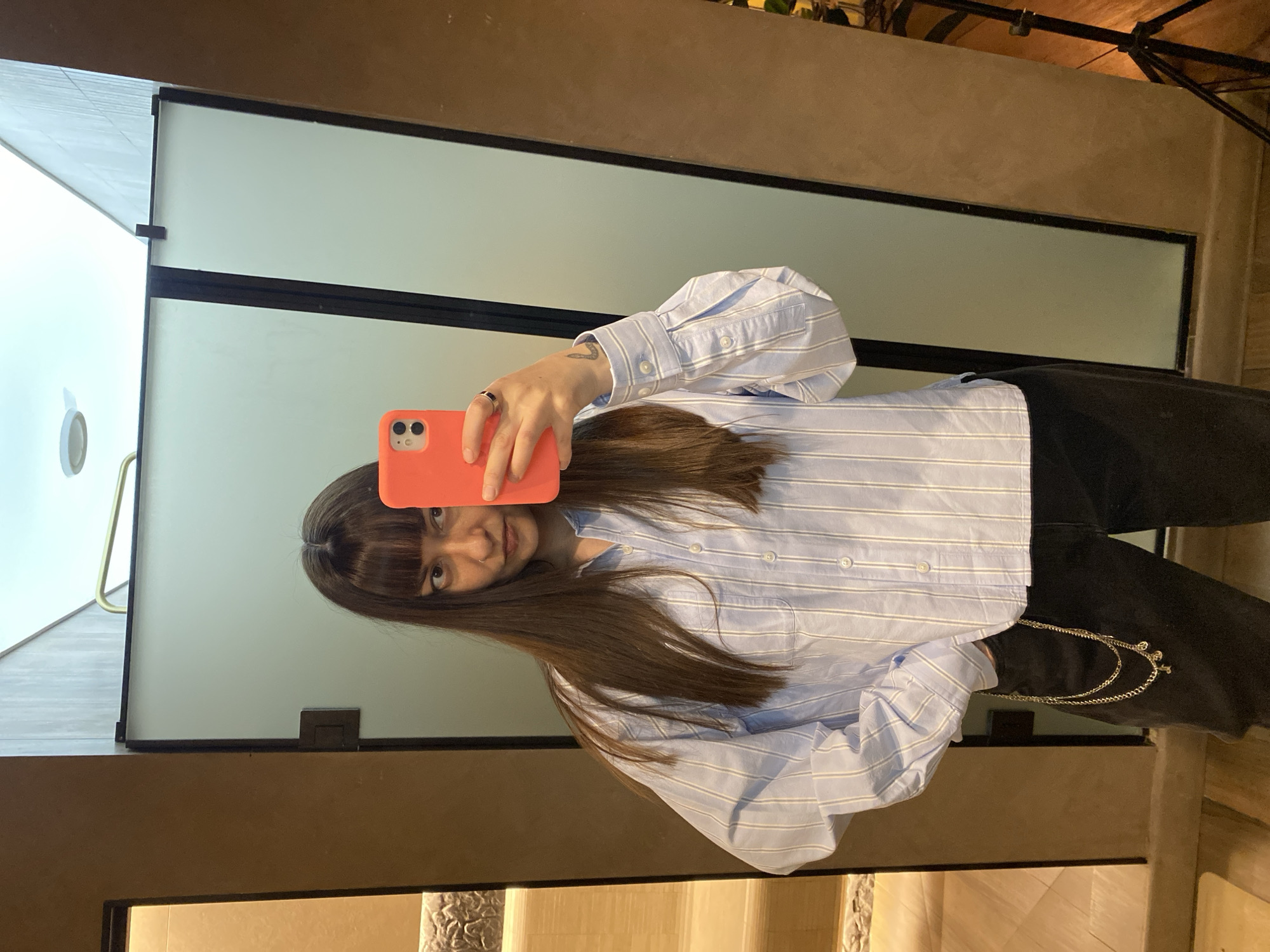 A woman with shoulder-length dark brown hair and bangs is taking a selfie in a mirror. She is holding an orange smartphone. She is wearing a light blue and white striped long-sleeved button-down shirt and black pants with a gold chain. The mirror appears to be part of a room divider or wall feature with black framing. The background includes a wall with a neutral color and what appears to be a staircase or decorative step. The environment suggests an indoor setting, possibly a residential or commercial space. There is no information about Izhevsk, Russia.