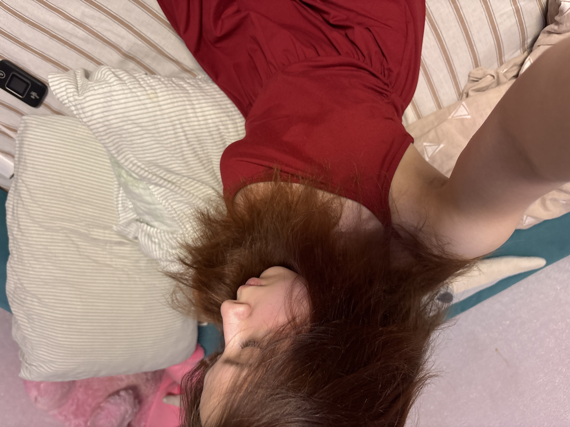 A woman lies on a bed with striped sheets and pillows. She wears a red dress. Her head is on a striped pillow, and her reddish-brown hair is spread around her face. A remote control is visible on the bed. The location provided is not evident in the image.