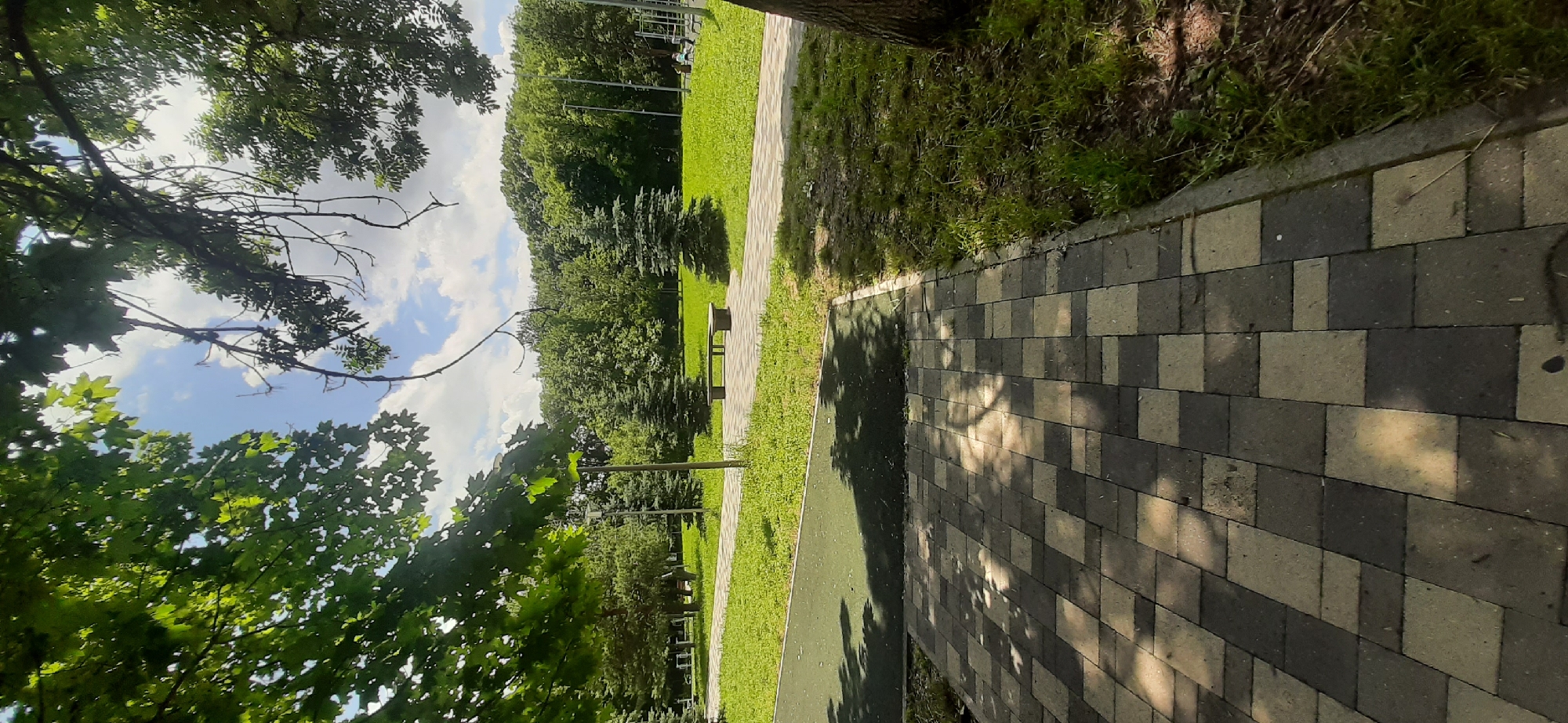The image shows a park path made of light and dark gray pavers, leading into a grassy area with trees and a bench. A green rubberized surface is visible in the foreground. In the background, there are various trees, including evergreen species, and a treeline of deciduous trees. The sky is partly cloudy. There are no people visible in the image. The overall scene depicts a public park area in Yessentukskaya, Russia.