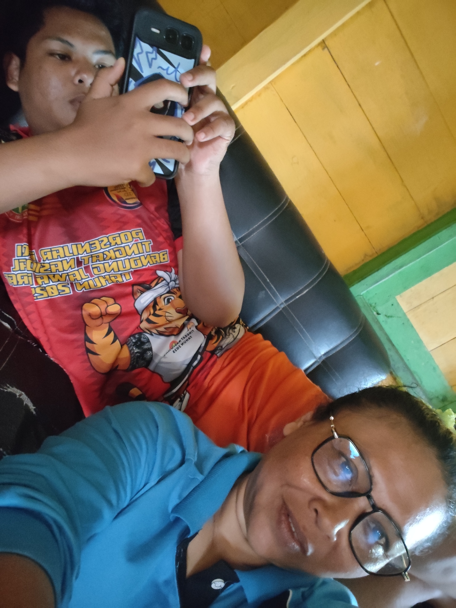 The image shows a close-up of two individuals, likely in a residential setting in Tanah Grogot, Indonesia. The person in the foreground is a woman wearing a blue shirt and eyeglasses, looking towards the camera. The person in the background is a young male holding a smartphone, wearing a red t-shirt with tiger graphics and text that appears to be in Indonesian. The background consists of a yellow wooden ceiling and a green painted wall, suggesting an indoor environment. The overall scene depicts a casual moment between two people.