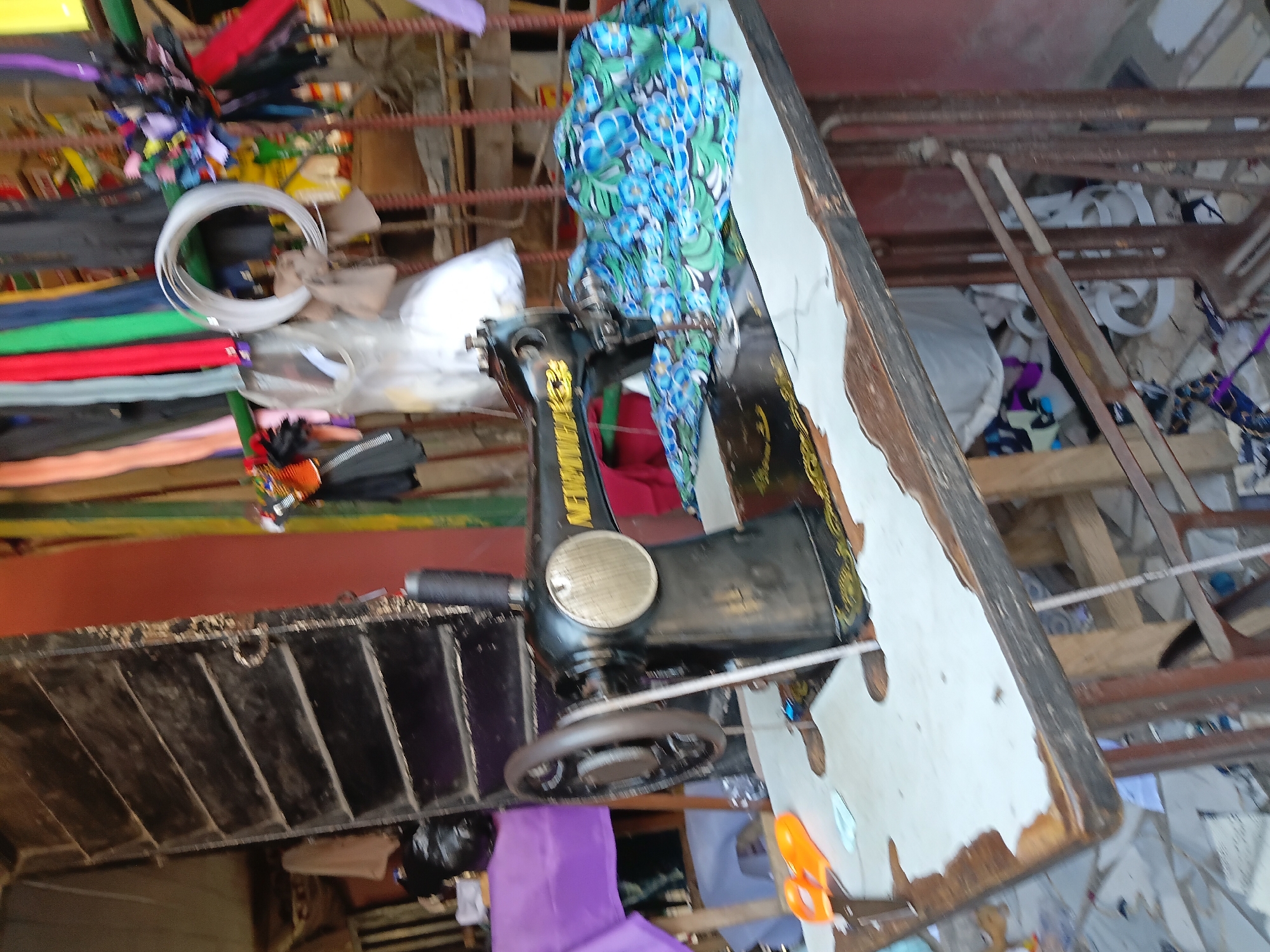 The image shows a sewing machine on a worn table, with fabrics and crafting supplies nearby. The setting appears to be a small shop in Ado, Nigeria, possibly a tailor's or seamstress's workspace, given the materials. The scene is cluttered, and the focus is on the sewing machine and its surrounding tools.