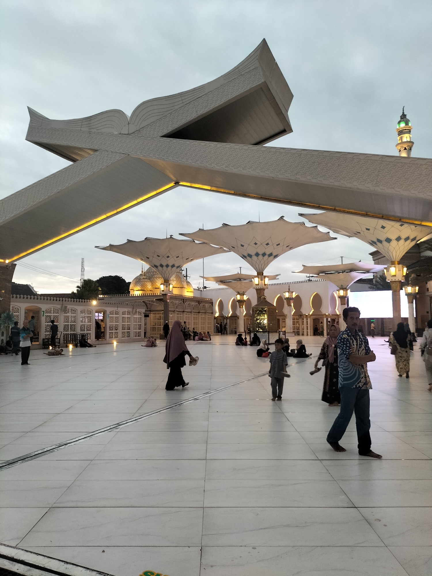 The image shows the courtyard of the Grand Mosque of Sheikh Zayed in the city of Blitar, Indonesia. The mosque features a large, white, canopy-like structure overhead, adorned with intricate patterns. Several large, white umbrellas providing shade are also visible, supported by illuminated pillars. The ground is paved with large, light-colored tiles.
In the foreground, a man in a patterned blue and white shirt and dark pants is walking, facing slightly to the left. He has his arms crossed and is barefoot. To his left, a young boy in a gray shirt and pants is walking, also barefoot, carrying a pair of shoes. Behind them, a woman wearing a dark hijab and black garment is walking, carrying her shoes.
Further back, in the courtyard, several other people are present. Some are walking, while others are seated on the ground. A woman in a light-colored hijab and patterned dress is walking towards the right side of the image. A minaret with a green top is visible in the background, illuminated by lights. The sky is overcast and appears to be twilight or early morning. The overall scene suggests a public gathering or visitation at the mosque.