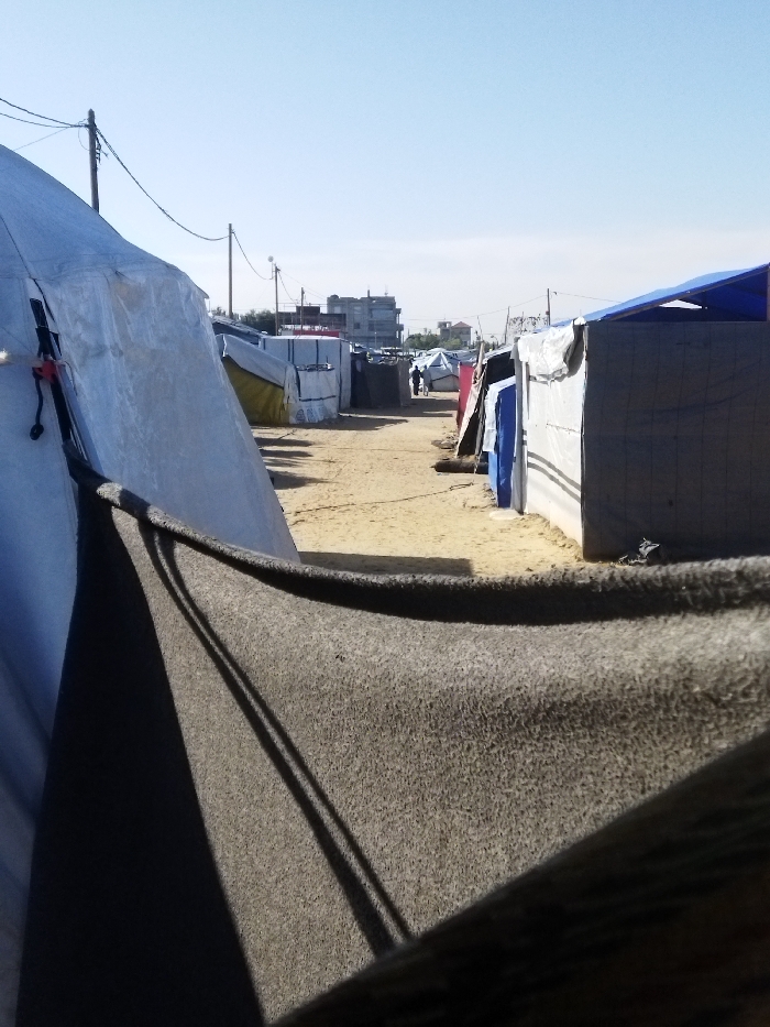 The image displays a ground-level view of a makeshift camp in Khan Yunis, Palestinian Territory, under a clear blue sky. A wide, sandy dirt path extends into the distance, flanked on both sides by numerous temporary shelters constructed from various materials including white, blue, and darker tarpaulins. In the immediate foreground, a dark grey, textured fabric, likely a blanket or tarp, hangs downward, partially obscuring the lower right view. To the left, a large tent made of white tarp material is visible, secured by dark ropes. Utility poles with overhead wires run along the path on both sides. In the midground, two indistinct figures are observed walking away from the viewer along the path. Multi-story buildings are visible in the background, situated beyond the camp structures.