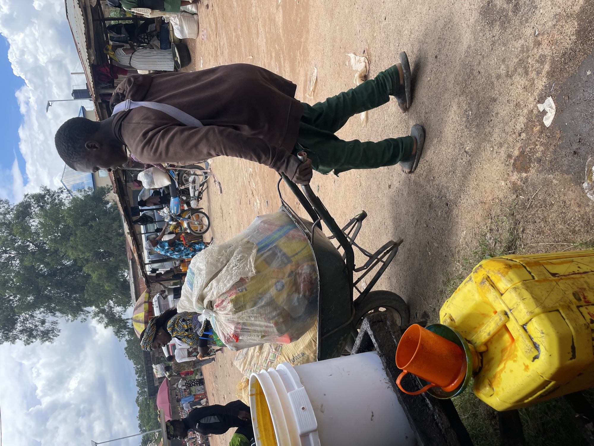 A man in a brown hoodie and green pants pushes a wheelbarrow loaded with large bags of goods. He is wearing sandals and has a strap across his chest. The wheelbarrow is also carrying a large white bucket with a yellow liquid inside. In the foreground, a yellow jerrycan with an orange cup on top is visible. The scene appears to be a marketplace with stalls and other people in the background. The ground is dirt. The location is identified as Wang, Nigeria.