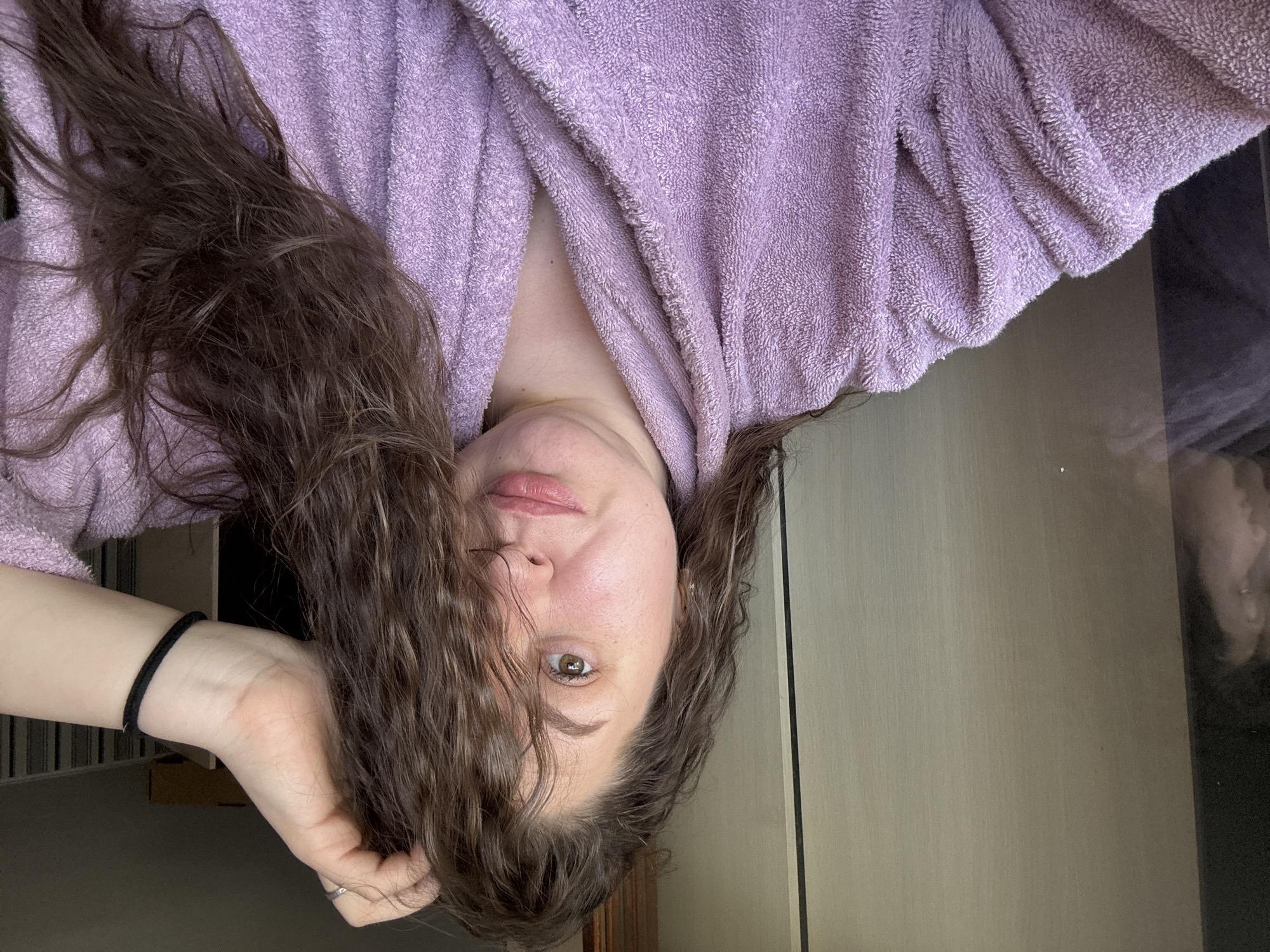 A woman is wearing a purple robe. Her hair is down and slightly obscures her face. Her hand is resting near her face, and she is looking directly at the camera. The background appears to be a room with light-colored walls. The location in the city of Марковский, Russia cannot be confirmed.