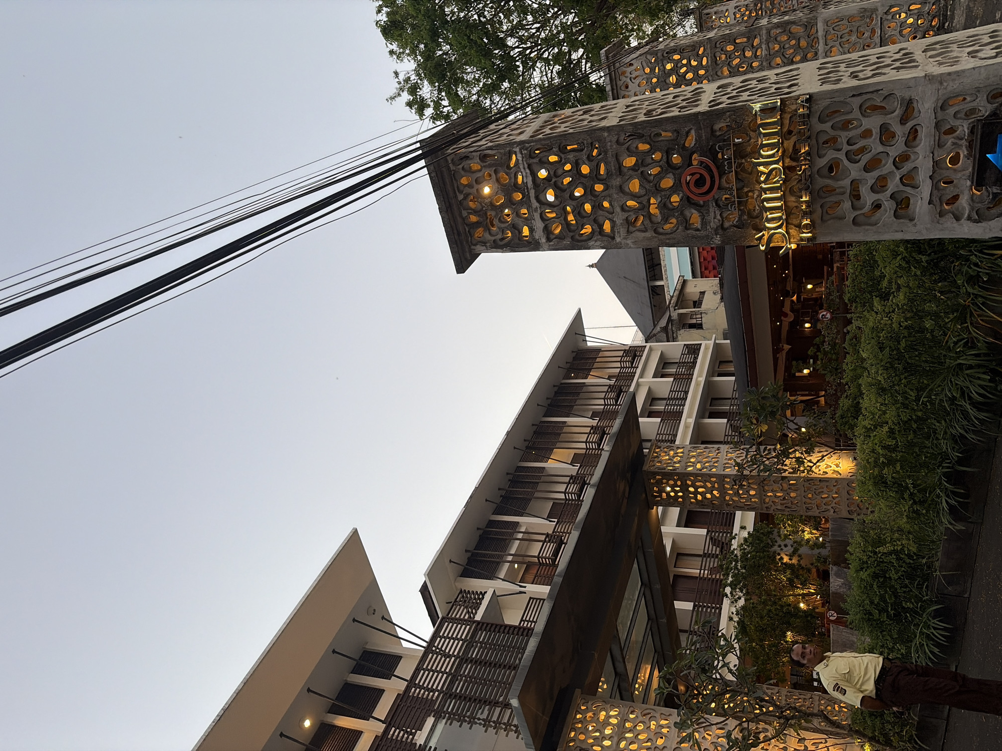 In Kuta, Indonesia, an outdoor shot captures the entrance to a hotel. A security guard in a light yellow uniform stands to the left of a decorative pillar with illuminated cutouts. Behind the guard, a modern hotel with multiple balconies and wooden accents is visible. Overhead, power lines crisscross against a pale sky. The hotel's name, "Samston Hotel & Spa," is illuminated in gold lettering on another pillar to the right. Lush green foliage surrounds the entrance.