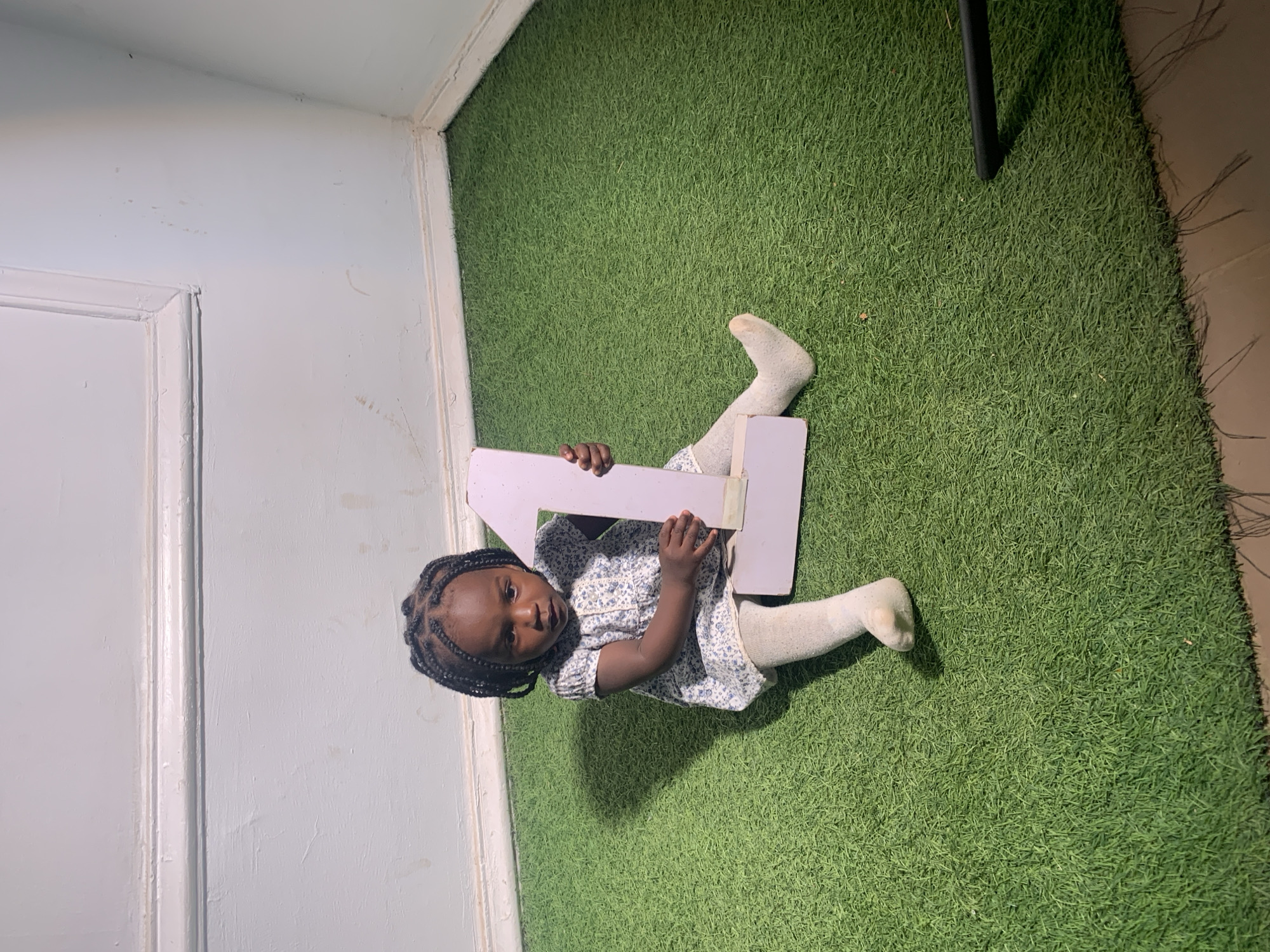 A young child sits on a green artificial turf. The child is holding a large wooden number "4". The background includes a white wall and door frame. It is not possible to determine the location in Barakin Akawo, Nigeria from the image.