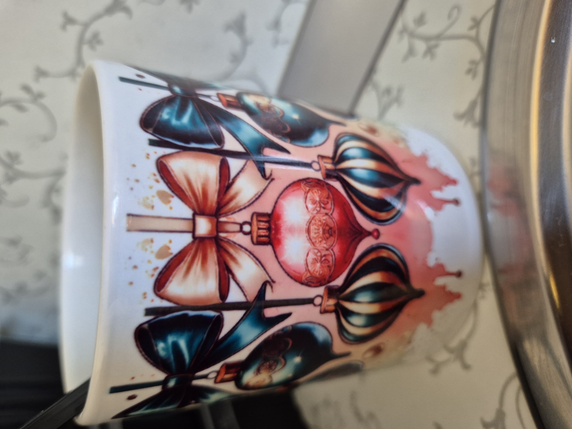 A close-up, eye-level shot shows a white ceramic mug decorated with a festive Christmas theme. The mug features a repeating pattern of ornate bows and hanging ornaments, rendered in shades of teal, coral, and gold. Red paint splatters are visible at the bottom of the mug, giving the impression of dripping liquid. A dark spoon or stirrer is partially visible inside the mug, and a black background and patterned wallpaper are visible behind it. The scene is set on a light-colored, patterned surface, with a reflective metal object in the foreground.