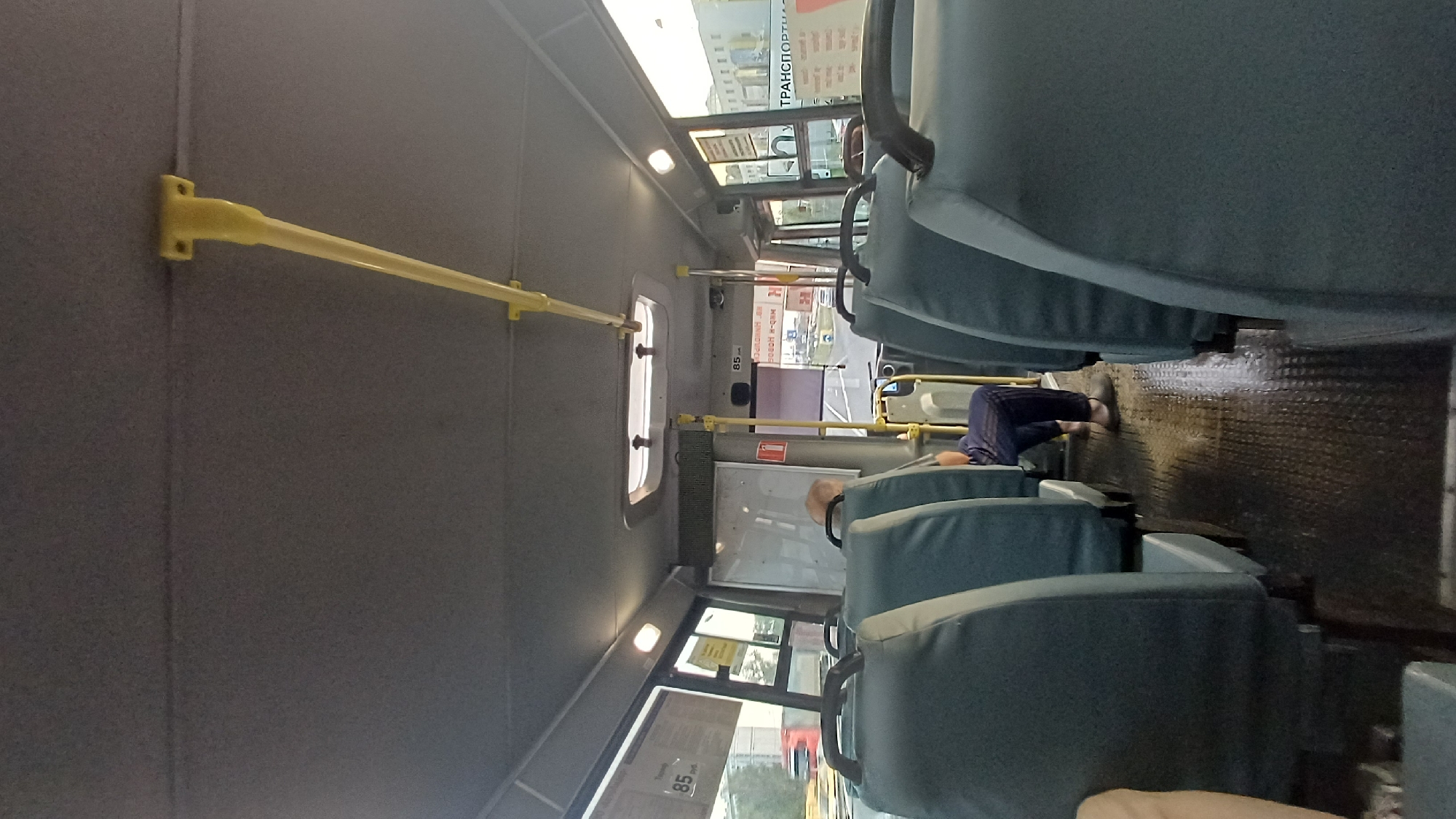 The image depicts the interior of a bus, likely in Balashikha, Russia. Multiple rows of grey fabric seats are visible. In the middle of the bus, a person wearing dark blue striped pants and grey slippers is seated, facing towards the back of the bus. The floor of the bus appears to be dark and textured, with some dampness or reflections visible on its surface, suggesting it may have recently rained or been cleaned. Yellow handrails are installed along the ceiling of the bus for passengers to hold onto. Through the windows, glimpses of a cityscape can be seen, including buildings and other vehicles. A white board or poster is mounted on the interior wall of the bus. A yellow bar is also visible attached to the ceiling.