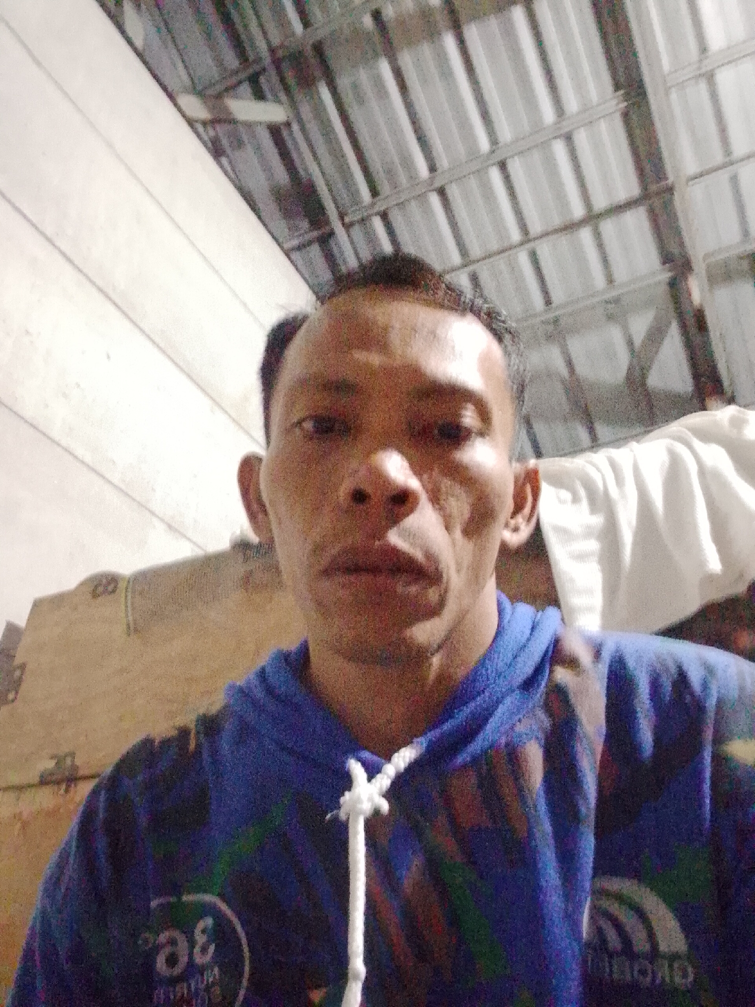 A man is indoors, likely in a residential dwelling in Bukit Ketok, Indonesia, as indicated by the metadata. He is wearing a blue hoodie with a white drawstring. The background includes corrugated metal roofing and possibly a bed with white sheets. The lighting is dim.