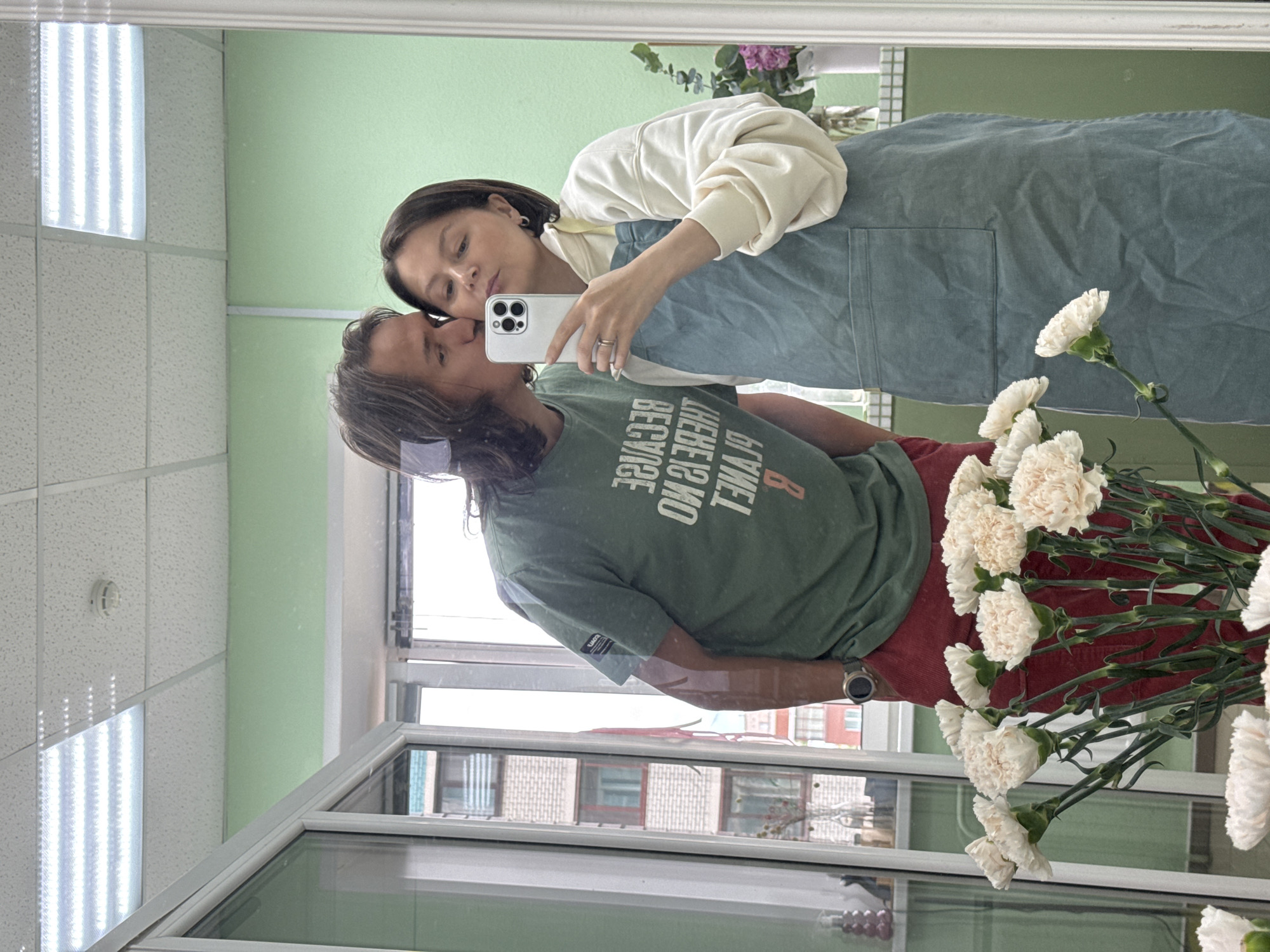 A woman is taking a selfie in a mirror with a man with long hair. The man is wearing a green t-shirt and red pants. The woman is wearing a white sweater and a blue apron. There are white carnations in the lower right of the frame. The reflection in the window shows a city landscape. Based on the architectural style of the buildings visible in the reflection, this may be Izhevsk, Russia.