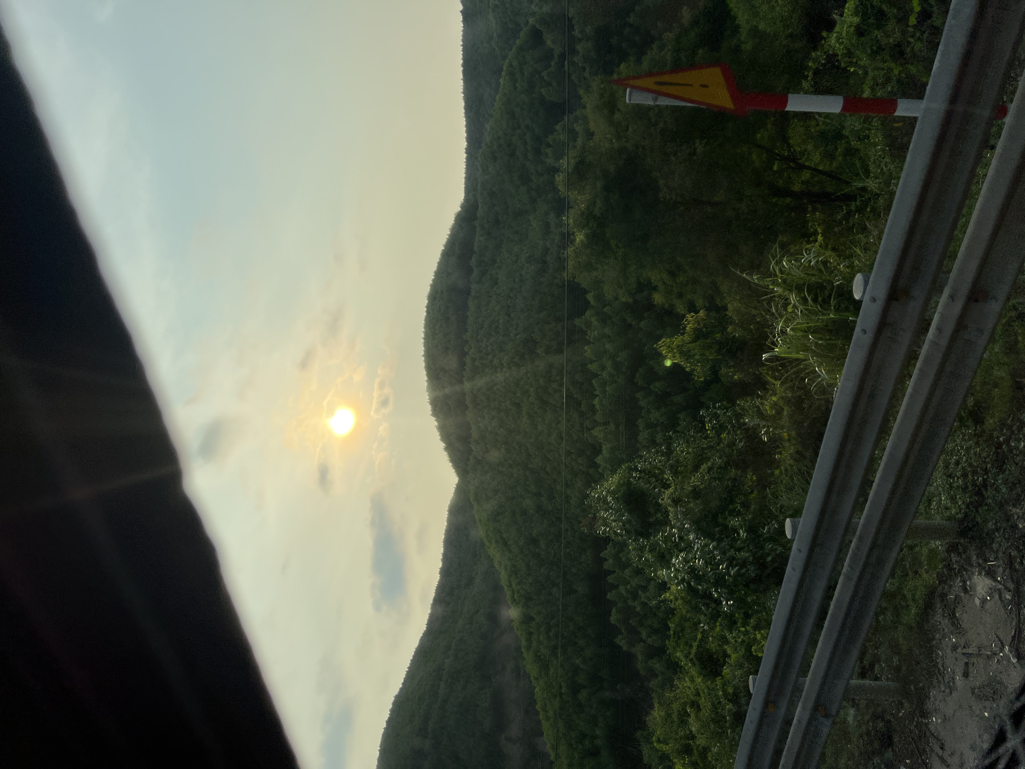 The image, taken from a moving vehicle, depicts a mountainous, forested landscape in Aê Thi, Vietnam. The sun is visible in the sky, partially obscured by clouds. A yellow and red road sign is positioned on the right side of the frame, near a metal guardrail. The guardrail is in the foreground, suggesting the image was taken from a road or highway. The mountain range extends into the distance, covered in dense vegetation. A portion of the vehicle's interior is visible on the left side of the frame.