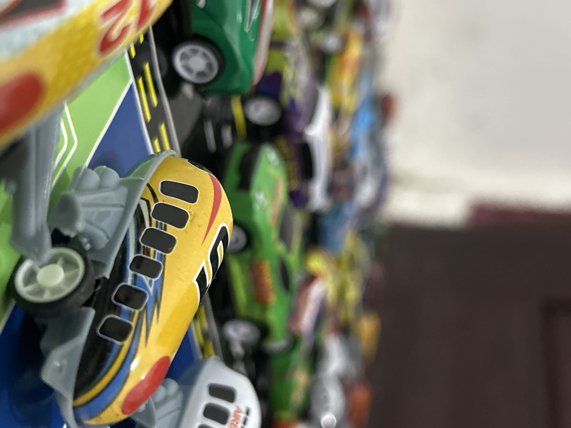 The image depicts a detailed close-up of a collection of miniature toy vehicles arranged on a multi-colored play surface, without any discernible geographic markers indicating the city of Lhokseumawe, Indonesia, or any other specific location.
In the foreground, sharply in focus, is a yellow toy designed to resemble a stylized bullet train or high-speed aircraft. It features black rectangular windows along its side, a blue and black striped pattern with a red nose cone, and is mounted on a grey plastic chassis with visible white wheels on the right side. Partially visible to the immediate left, another similar yellow toy is out of focus, showing red lettering. These foreground toys are positioned on a playmat or surface that features distinct sections of blue and green, bordered by black strips with yellow lines, mimicking road or track markings.
The midground, progressively less in focus, reveals numerous other toy cars arranged on similar black "road" sections. Identifiable vehicles include a dark green sports car on the left with white wheels, a purple and white striped car in the center, and a lime green car with yellow/orange lettering on the right. These cars exhibit varying designs and colors, suggesting a diverse collection.
The background is heavily blurred and extends upwards, showing more indistinct shapes of toy vehicles. The upper-left background appears as a light, uniform surface, possibly a wall, while the upper-right background is a darker, warm brown or maroon tone, indicating another indoor surface or piece of furniture. No people are visible in the scene. The lighting is soft and even, highlighting the vibrant colors of the toys.