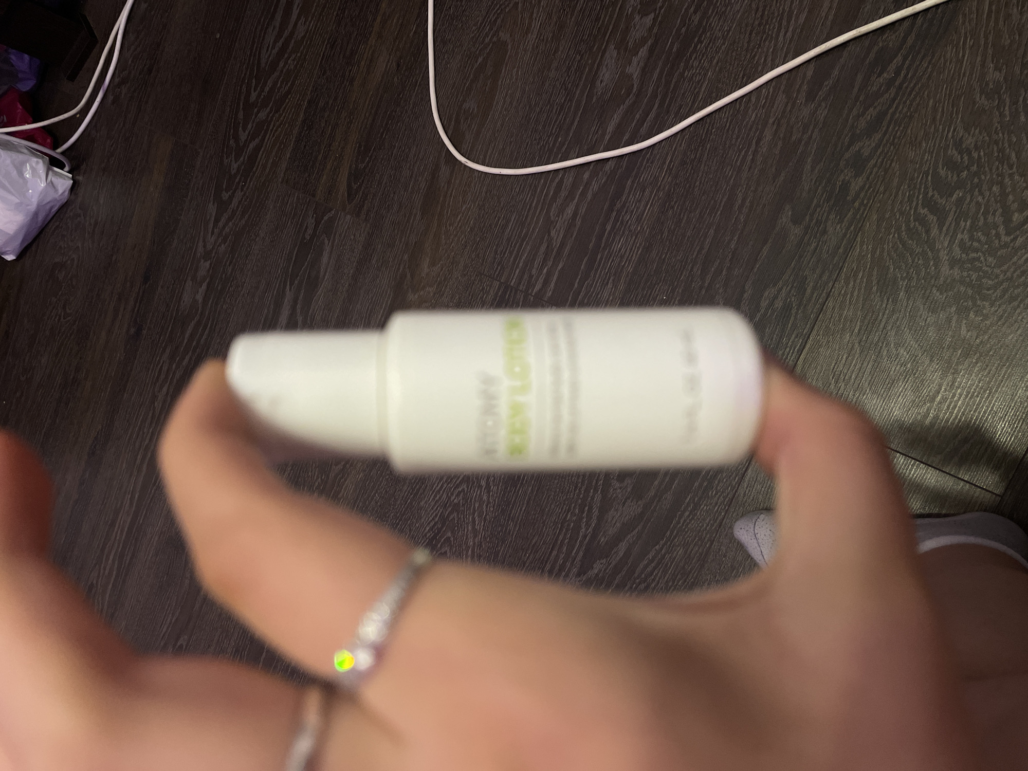 A close-up shot shows a hand holding a small white bottle of body lotion. The bottle has green text that is partially legible, but appears to say "BODY LOTION". A portion of a ring with a small, iridescent green and yellow stone is visible on one of the fingers. The background is a dark wooden floor. A white cable snakes across the floor in the upper right corner of the image, and some white and purple packaging is visible further in the background. The scene is not identifiable as being in Shcheglovo, Russia.
