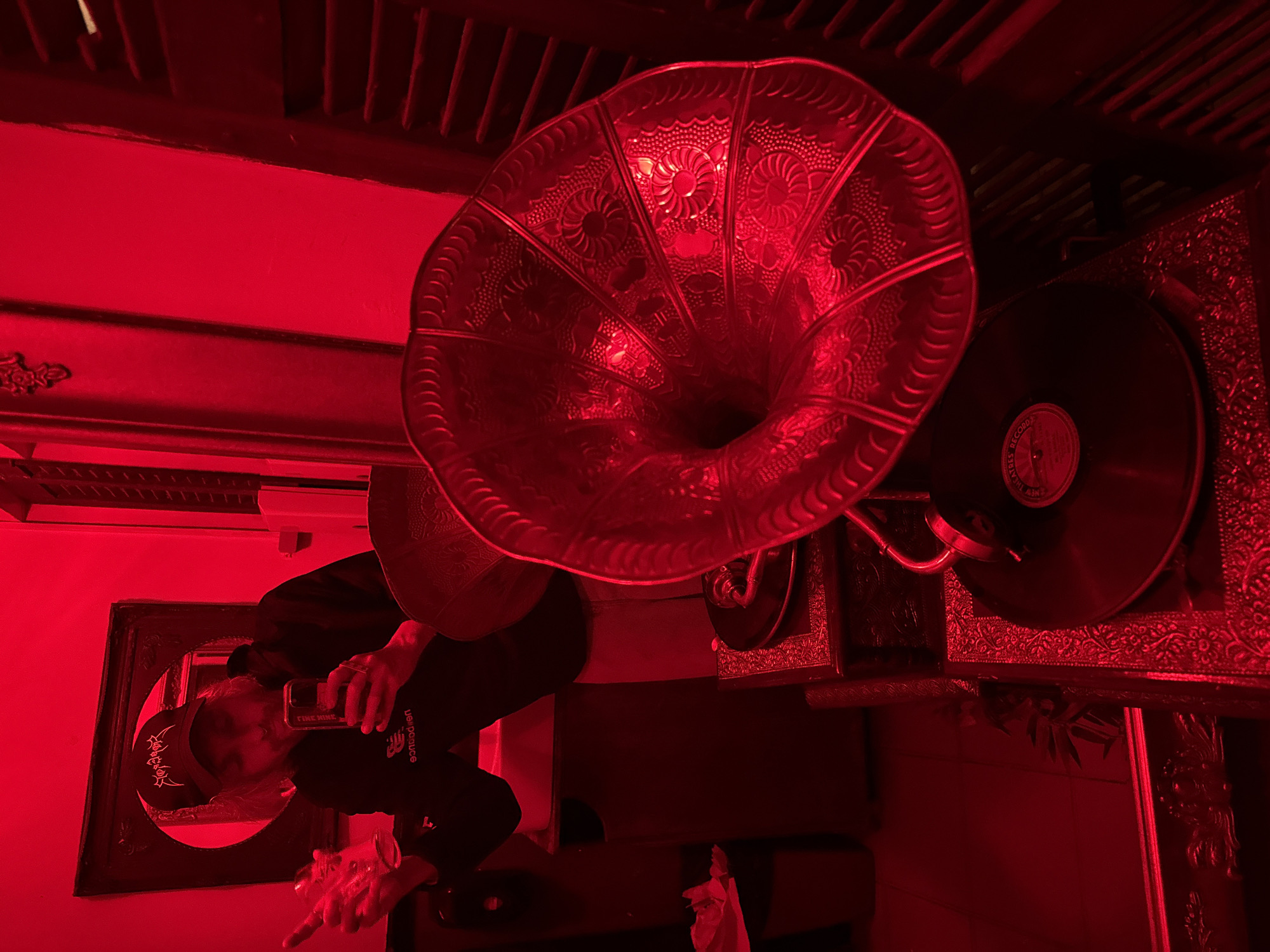 A red-lit room features a vintage gramophone with a large, ornate horn and a vinyl record. A person is visible in a mirror, wearing a baseball cap and holding a mobile phone. The location is possibly in Tel-Aviv, Israel, although this is not directly evident in the image.