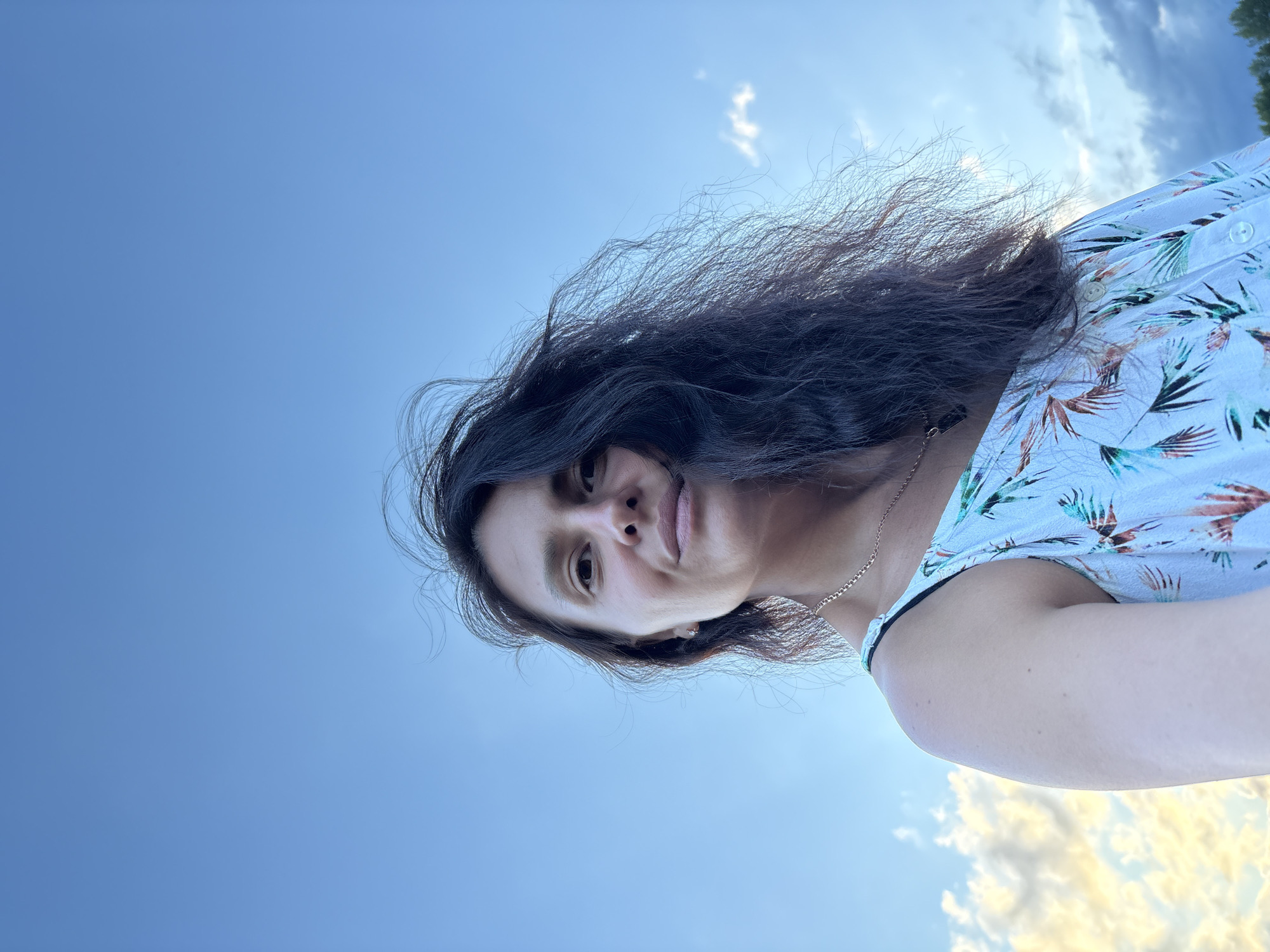 A low-angle, close-up, outdoor, selfie-style photograph features a woman with long, dark, wavy hair against a bright blue sky with scattered clouds. She is wearing a light-colored, sleeveless dress with a tropical leaf pattern. Her head is tilted slightly, and she is looking towards the camera with a subtle smile. The background is dominated by the sky, with a hint of clouds tinged with yellow light at the bottom. The image appears to have been taken in Chaikovsky, Russia, given the context provided.