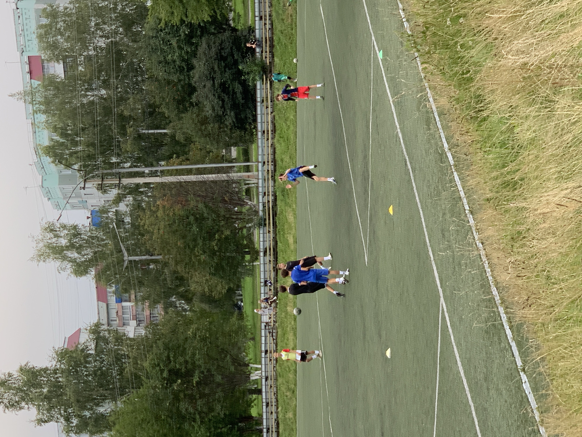 A group of young males are playing soccer on an outdoor artificial turf field. The field is marked with white lines and equipped with yellow and green cones. In the background, several trees and residential buildings are visible, indicating an urban setting. The sky is overcast. The people are dressed in athletic clothing, and their actions suggest a game or practice session is underway. The overall scene is consistent with a recreational area within the city of Prokopyevsk, Russia.