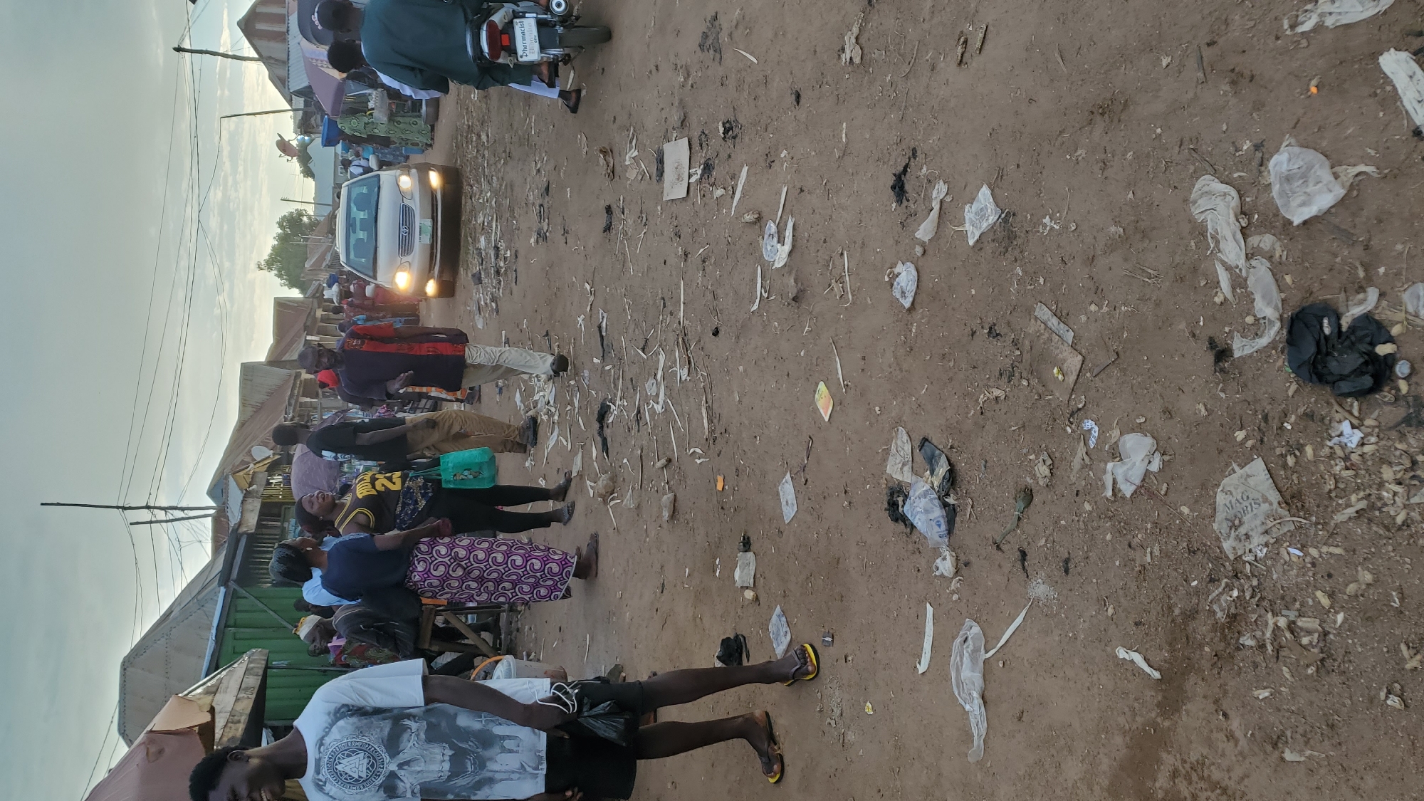A group of people are seen walking on a dirt road in Jalingo, Nigeria. The road is littered with debris, including plastic bags and wrappers. Several market stalls with corrugated iron roofs are visible on either side of the road. A silver car is driving down the road, and a motorcycle is parked on the right side. The sky is overcast. The people are dressed in casual clothing, and some appear to be engaged in conversation. The overall scene depicts a busy market environment with signs of litter.